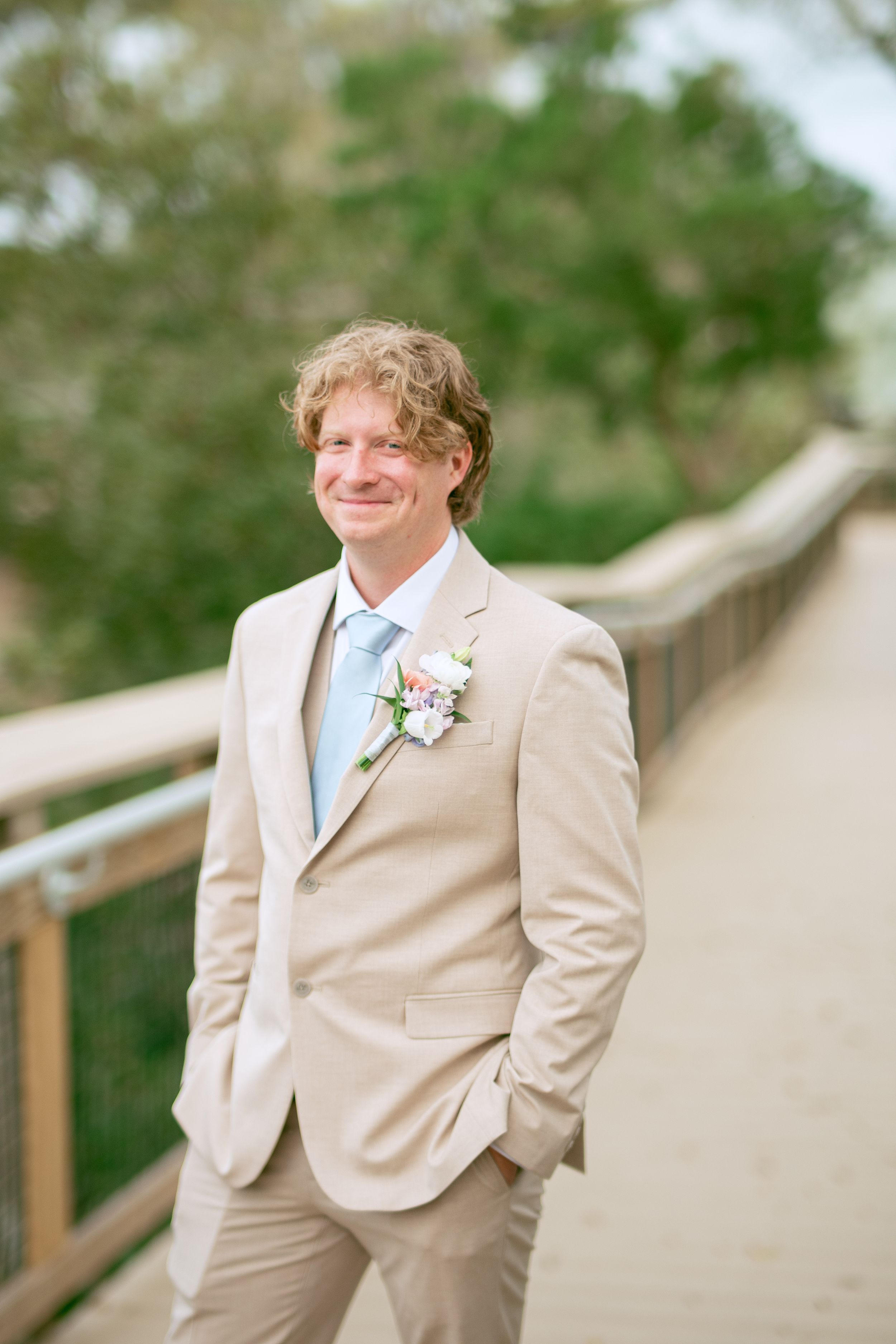 A groom in a light beige suit with blue tie and boutonniere poses on a wooden walkway.