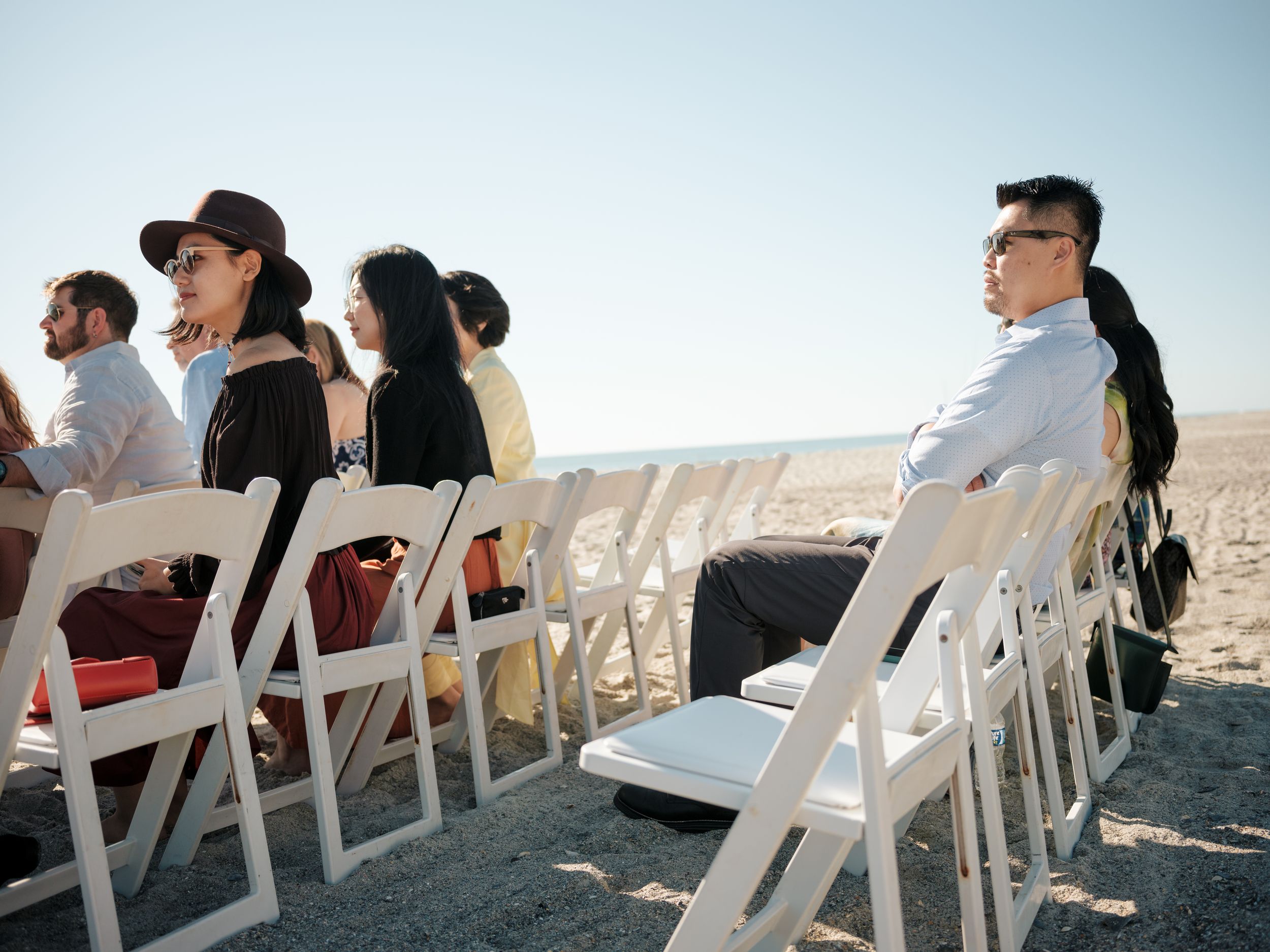Row of wedding attendees watching beachside ceremony from white folding chairs.
