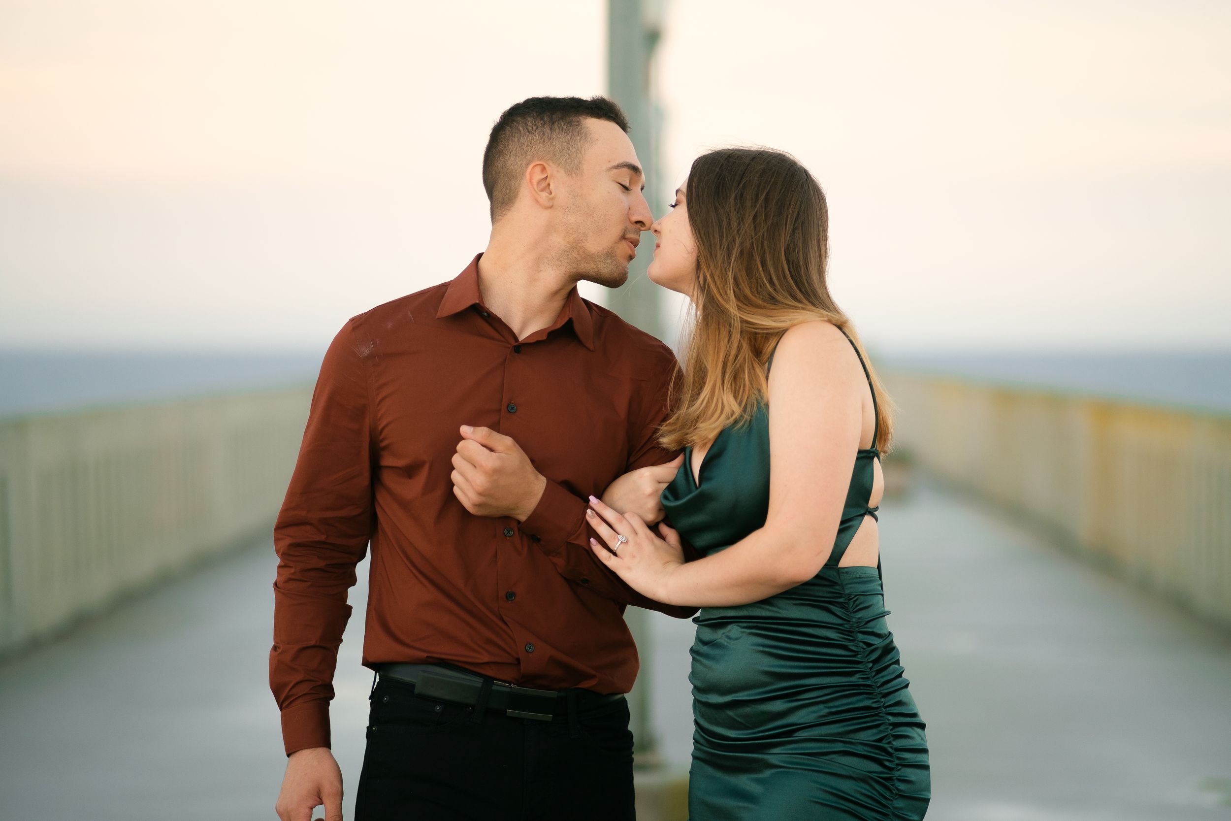 A romantic couple in formal attire sharing an intimate moment on a pier at sunset.