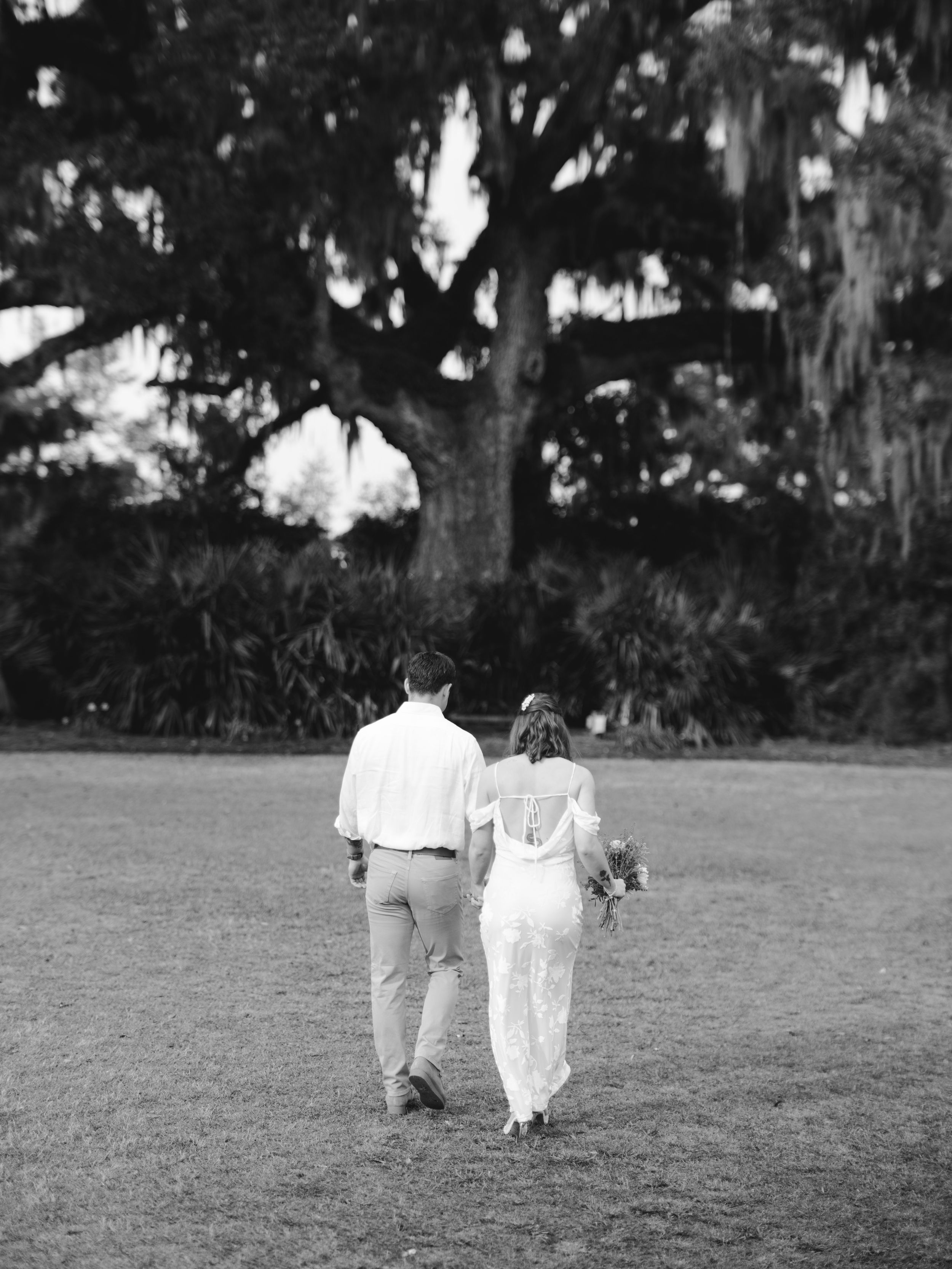 A couple wearing white outfits walks hand in hand across an outdoor grassy area in black and white.