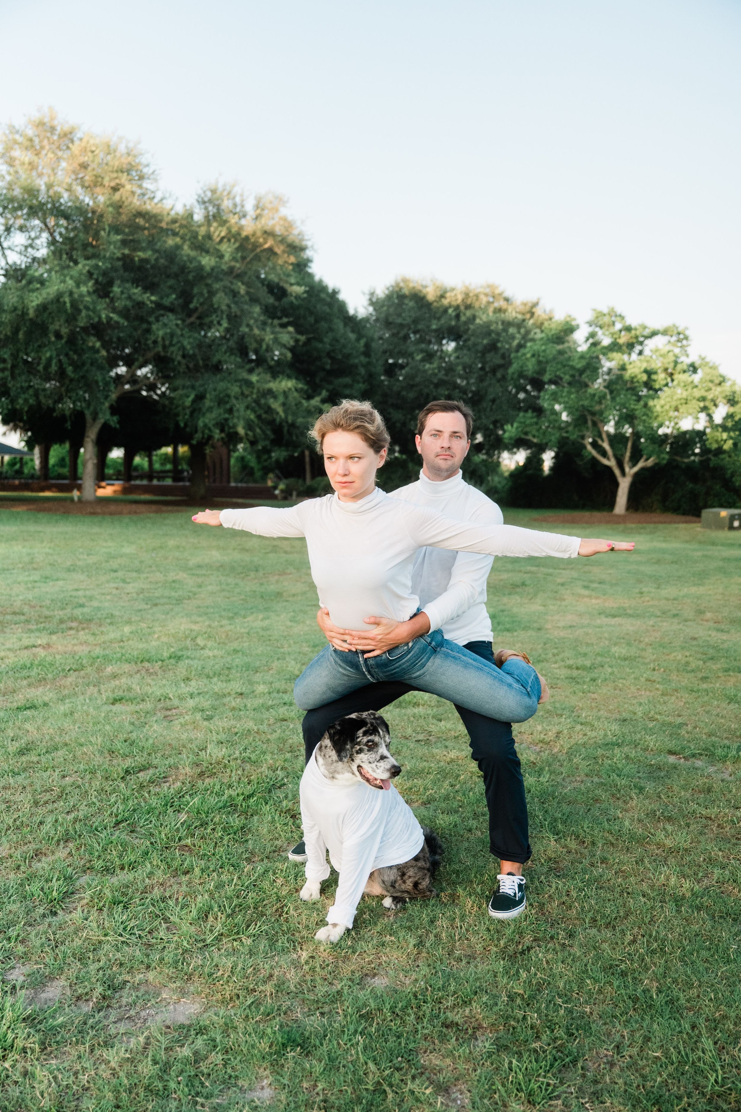 A couple poses playfully in a grassy park with their Australian Shepherd dog on a sunny afternoon.