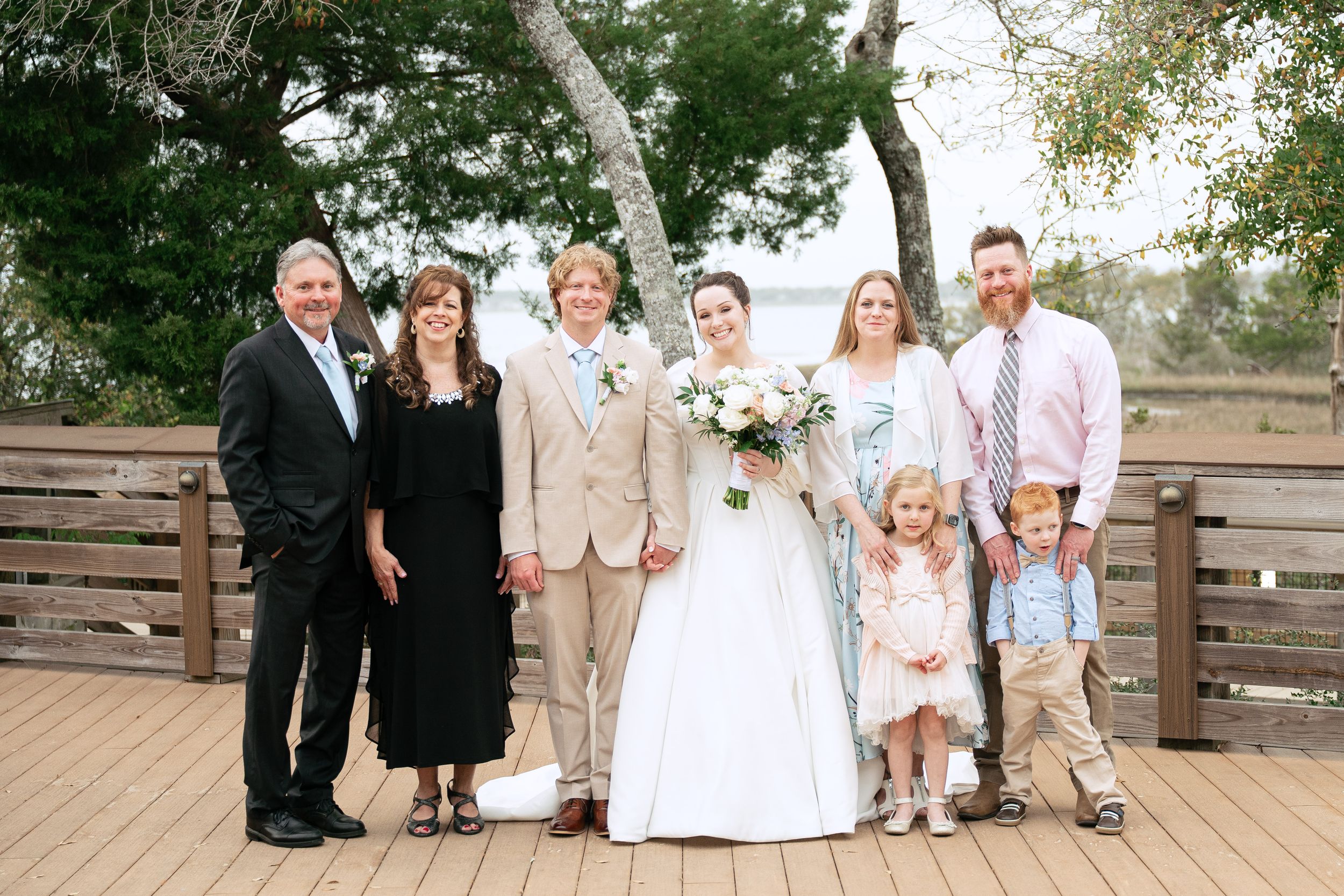 A wedding party poses together on a wooden deck outdoors for a formal group photo.