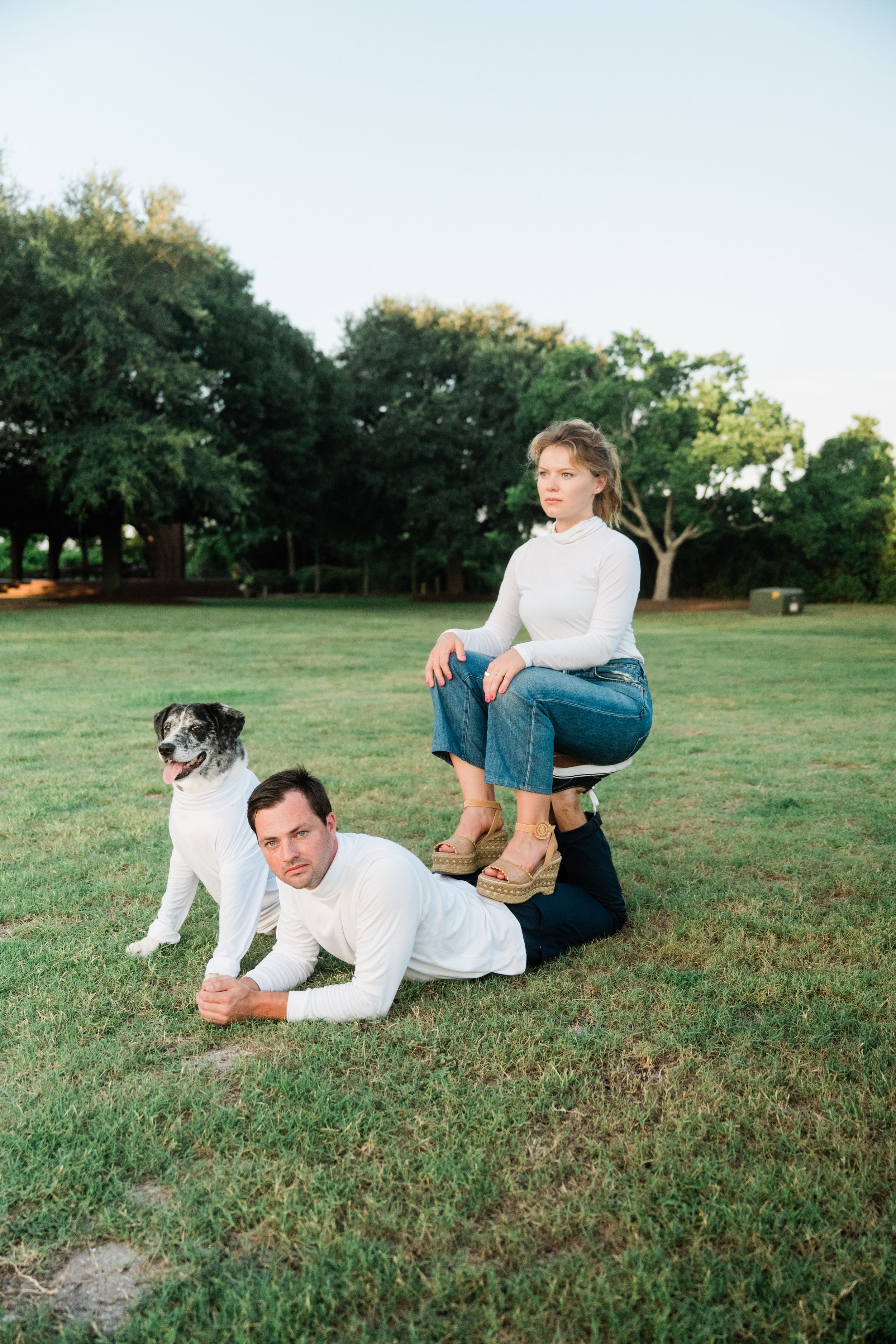 Playful sequence of people lying and rolling on grassy field with dog in casual white outfits at sunset.