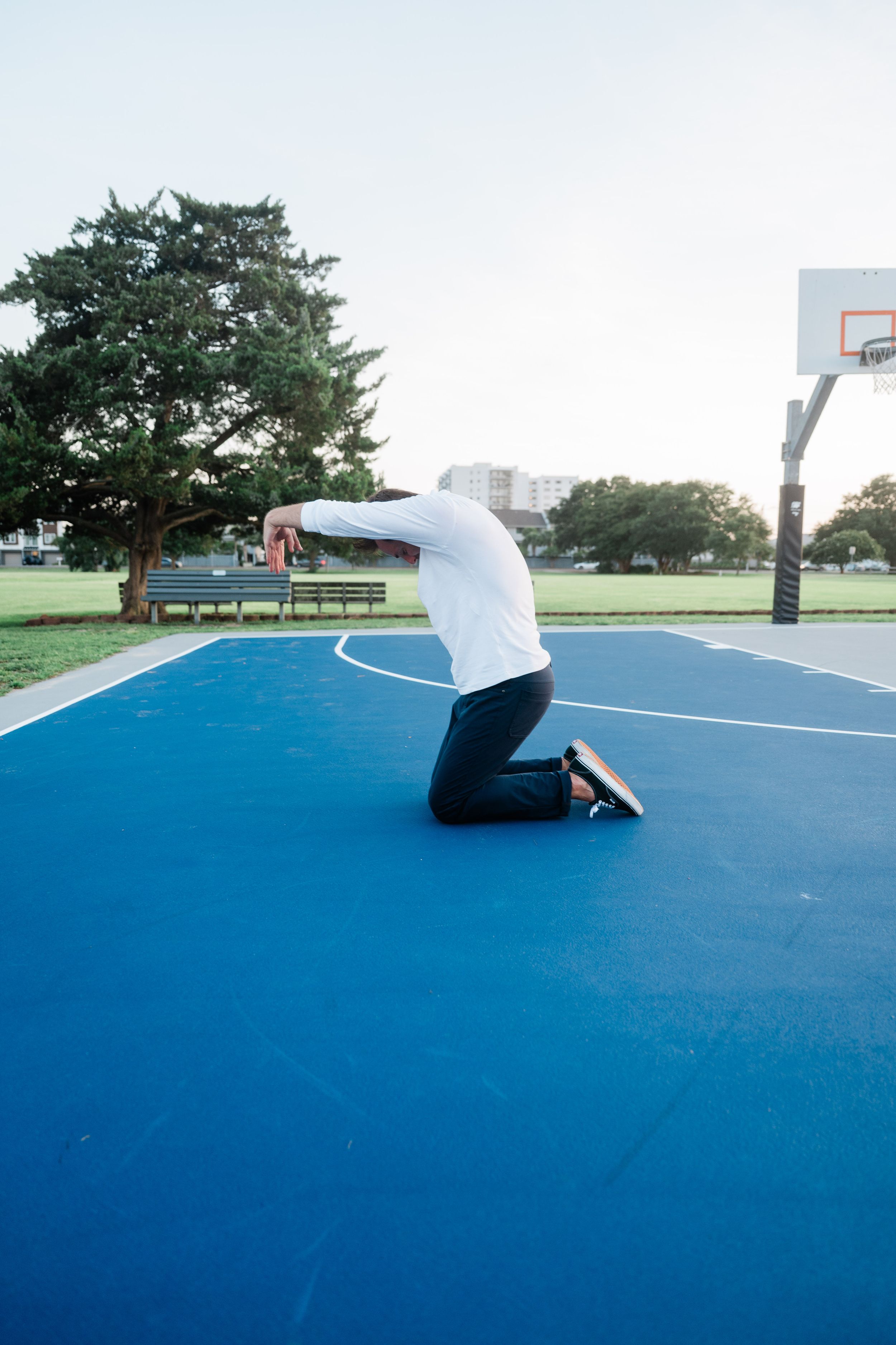 A person falls to their knees on a bright blue outdoor basketball court with trees and a hoop in the background.