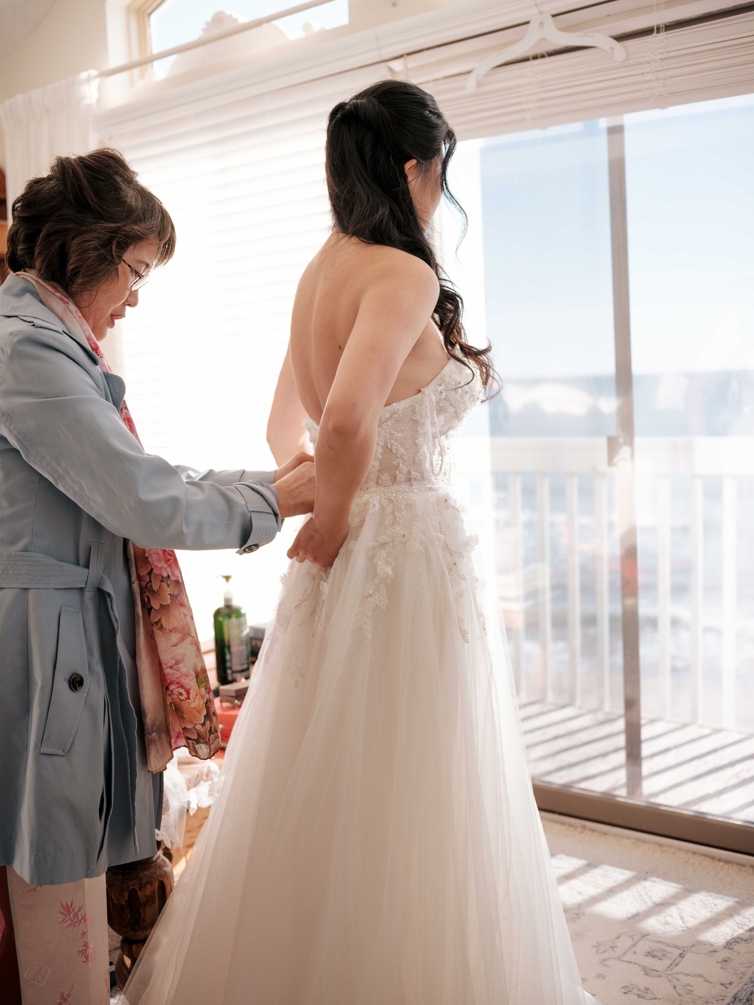 A flowing white wedding dress being adjusted in a sunlit room with city views.