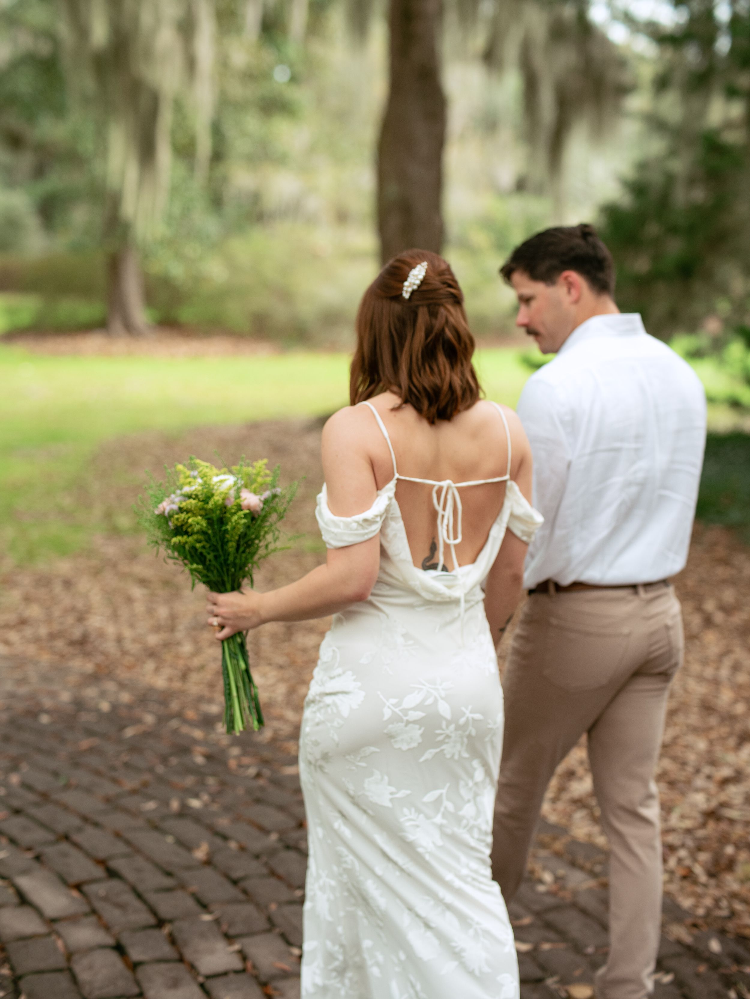 A couple in wedding attire stroll along a brick path surrounded by lush garden scenery.