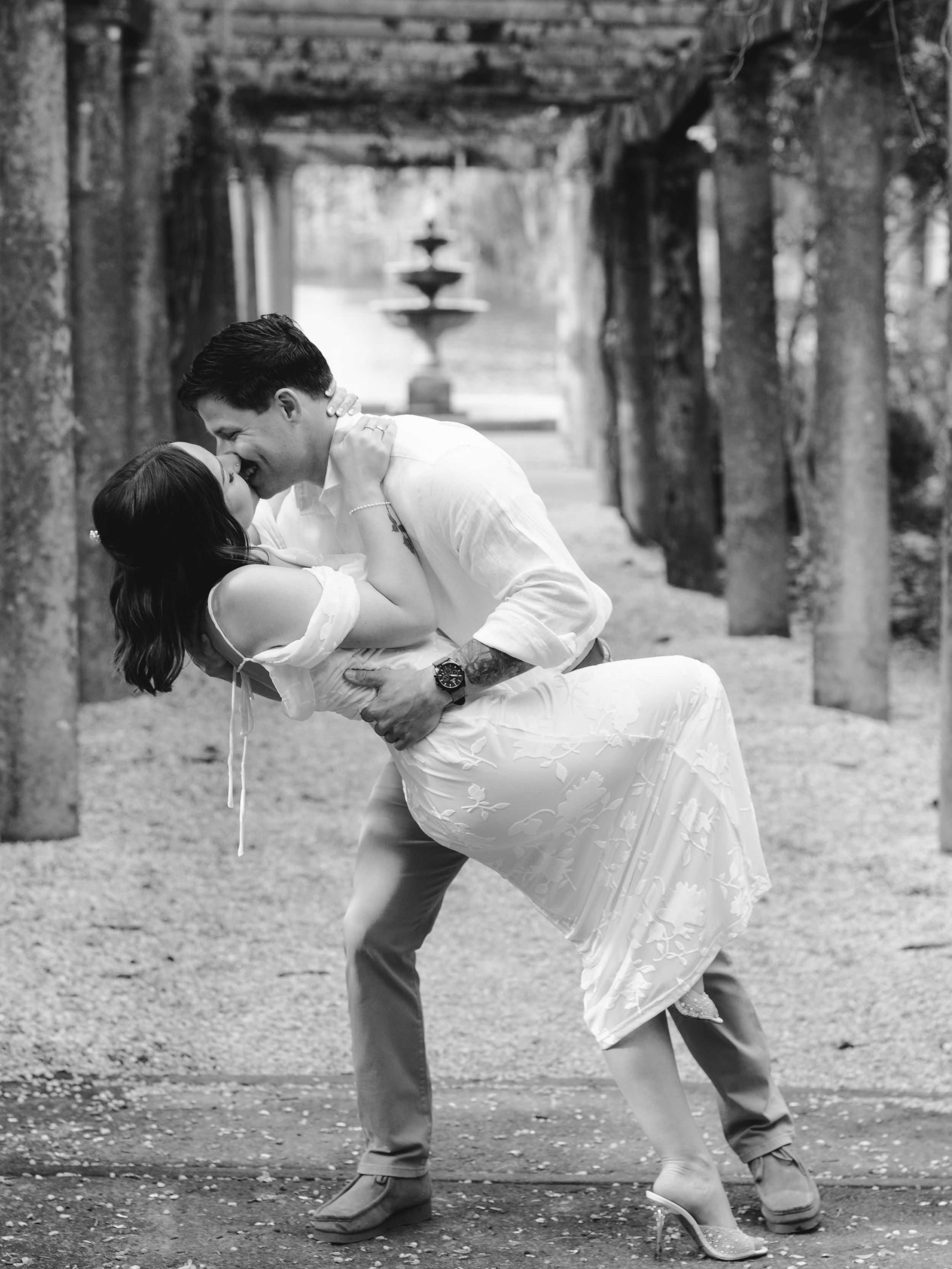 A romantic black and white photo of a couple in wedding attire sharing a passionate dip kiss in front of a fountain.