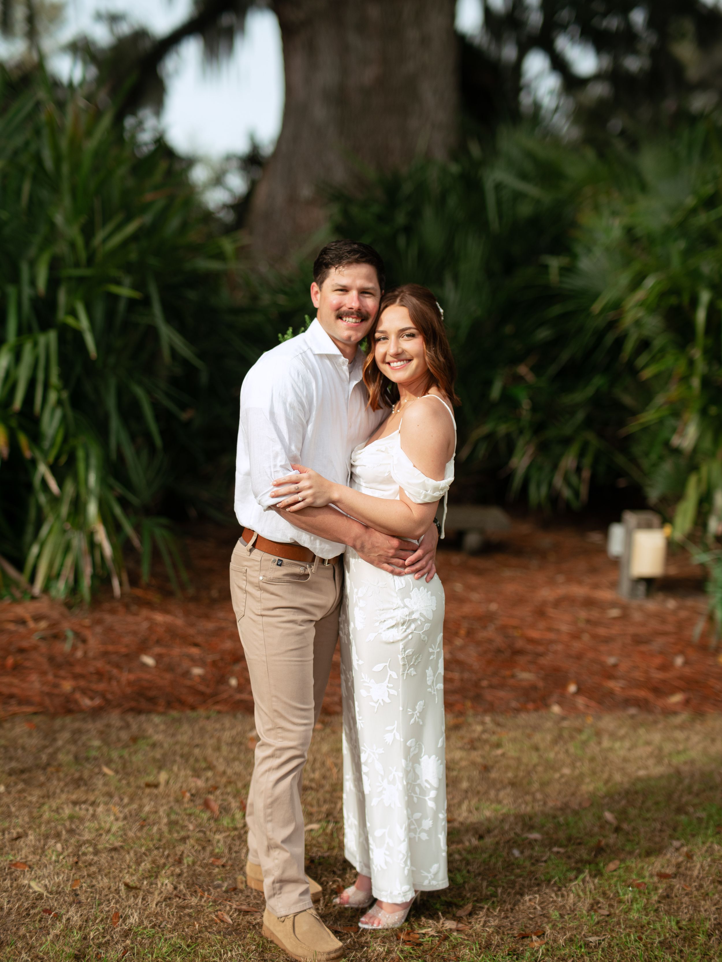 A romantic couple in casual attire embraces while standing on mulch surrounded by tropical plants.