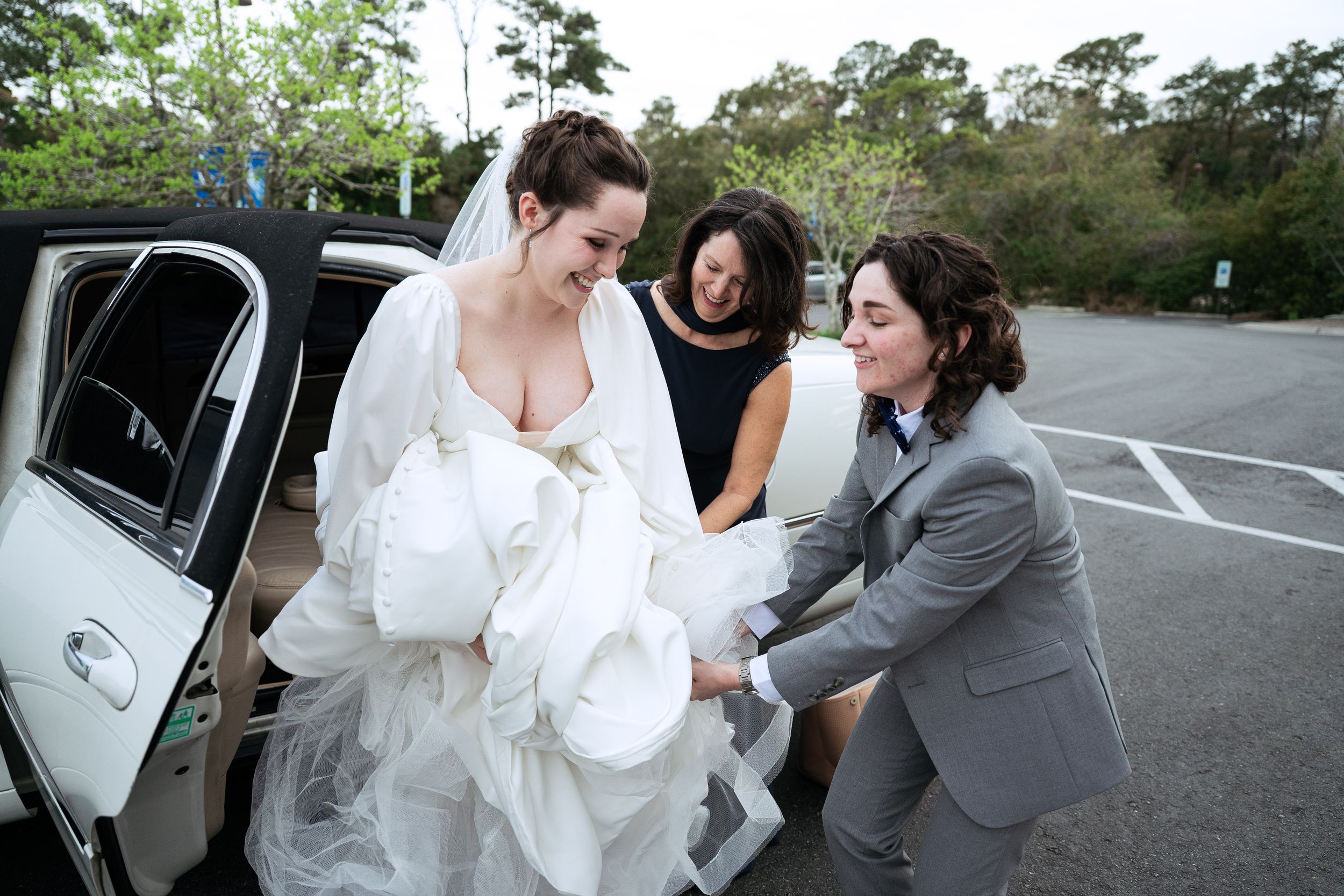 A wedding party helps the bride into a white van in a parking lot.