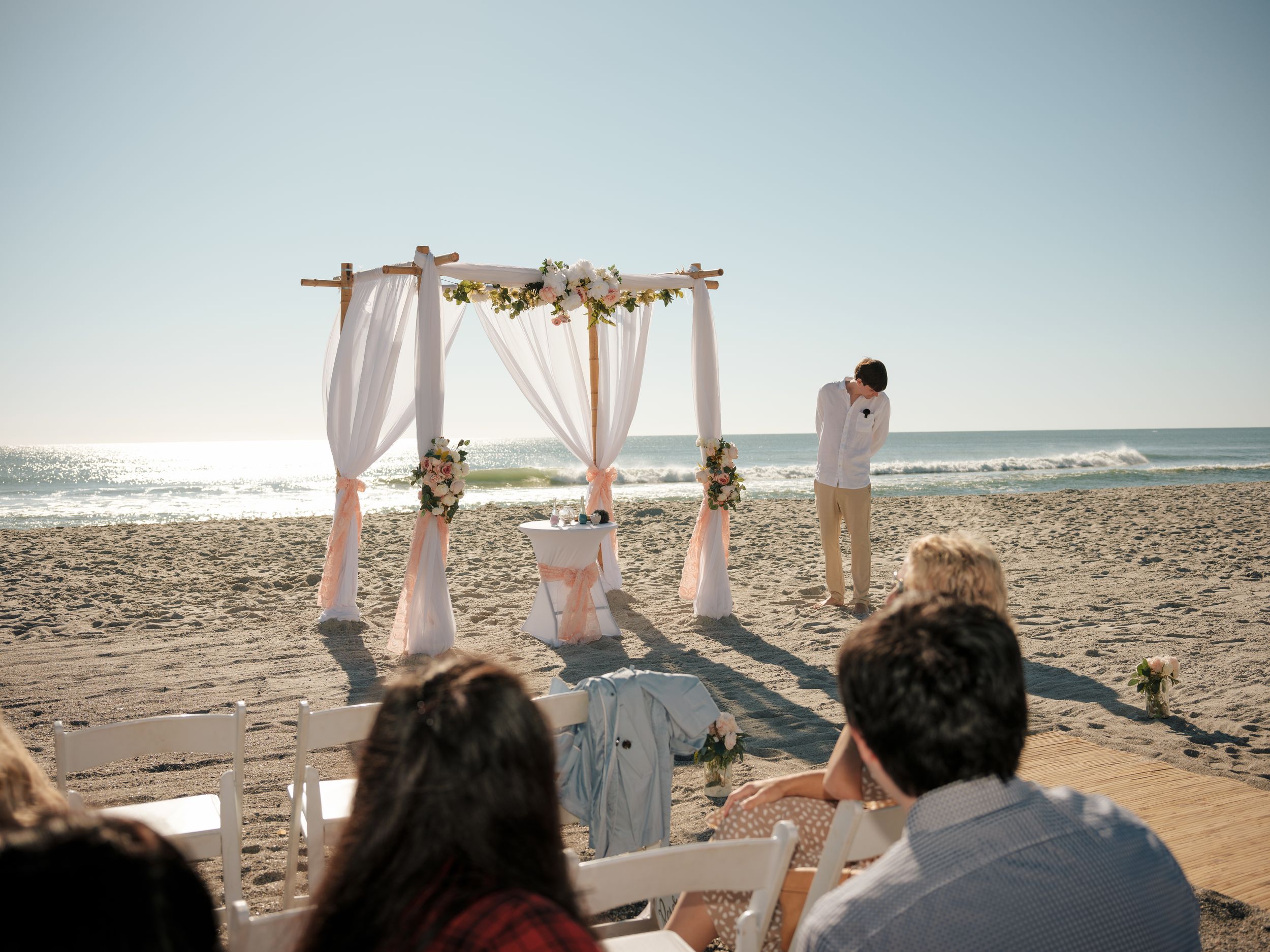 Wedding guests seated in white chairs on a sandy beach for an oceanfront ceremony.