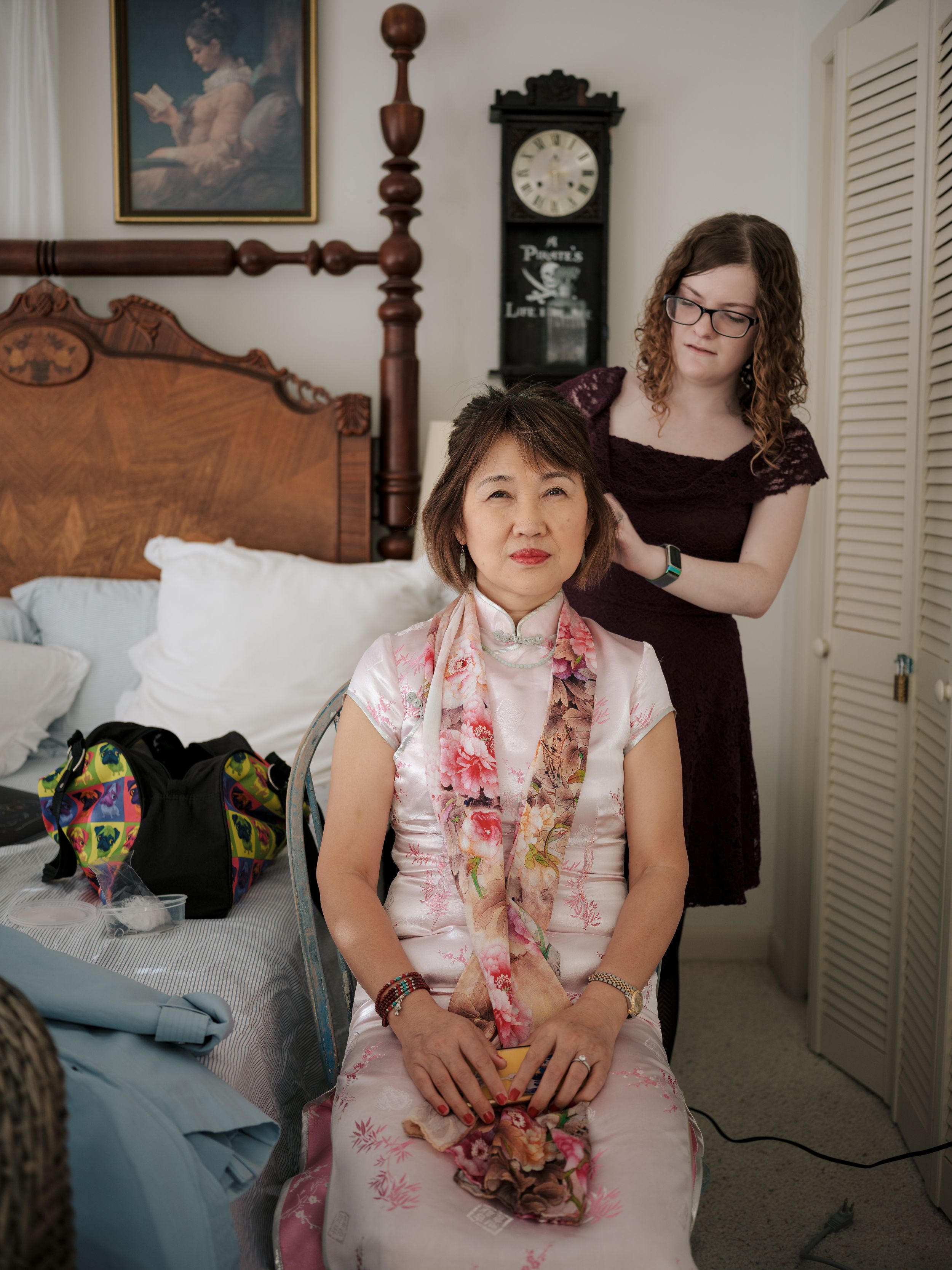 Person in white and pink dress sits on decorative wooden bed against a beige wall with traditional style furniture.