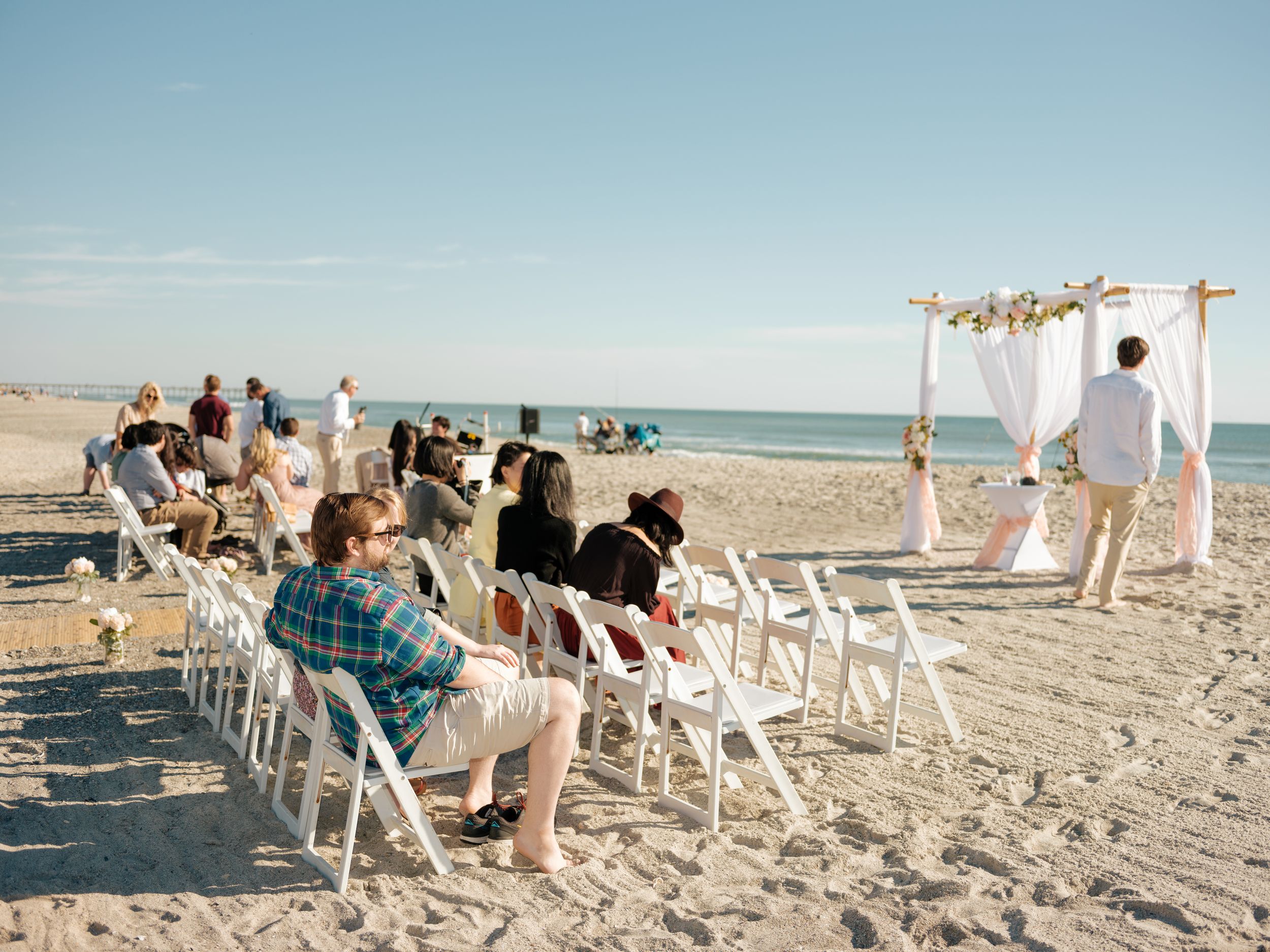 Beach wedding ceremony setup with white chairs and floral arch against the ocean backdrop.