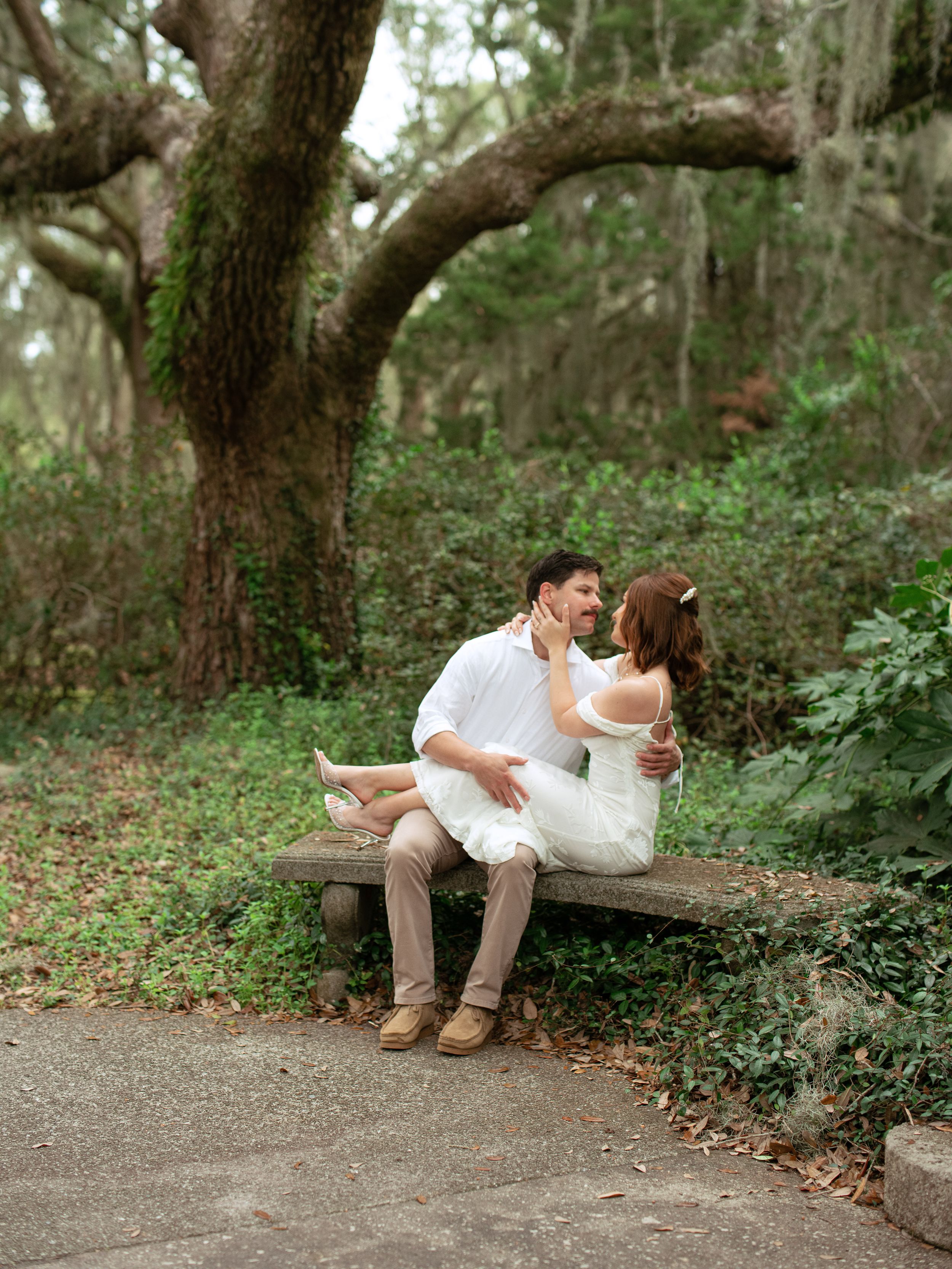 A couple lounges together on a rustic park bench surrounded by lush greenery and trees in a romantic outdoor setting.