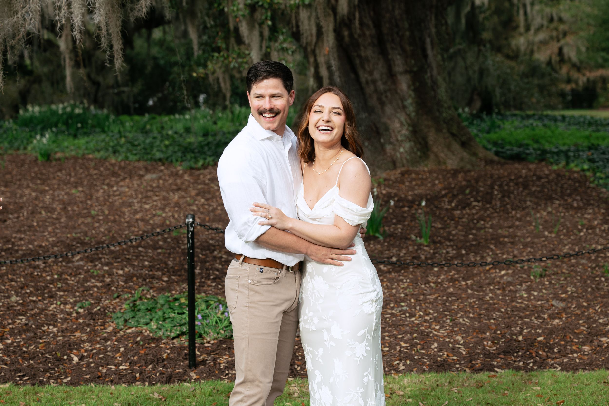 A couple in white and beige clothing embraces while taking outdoor photos in a garden setting.