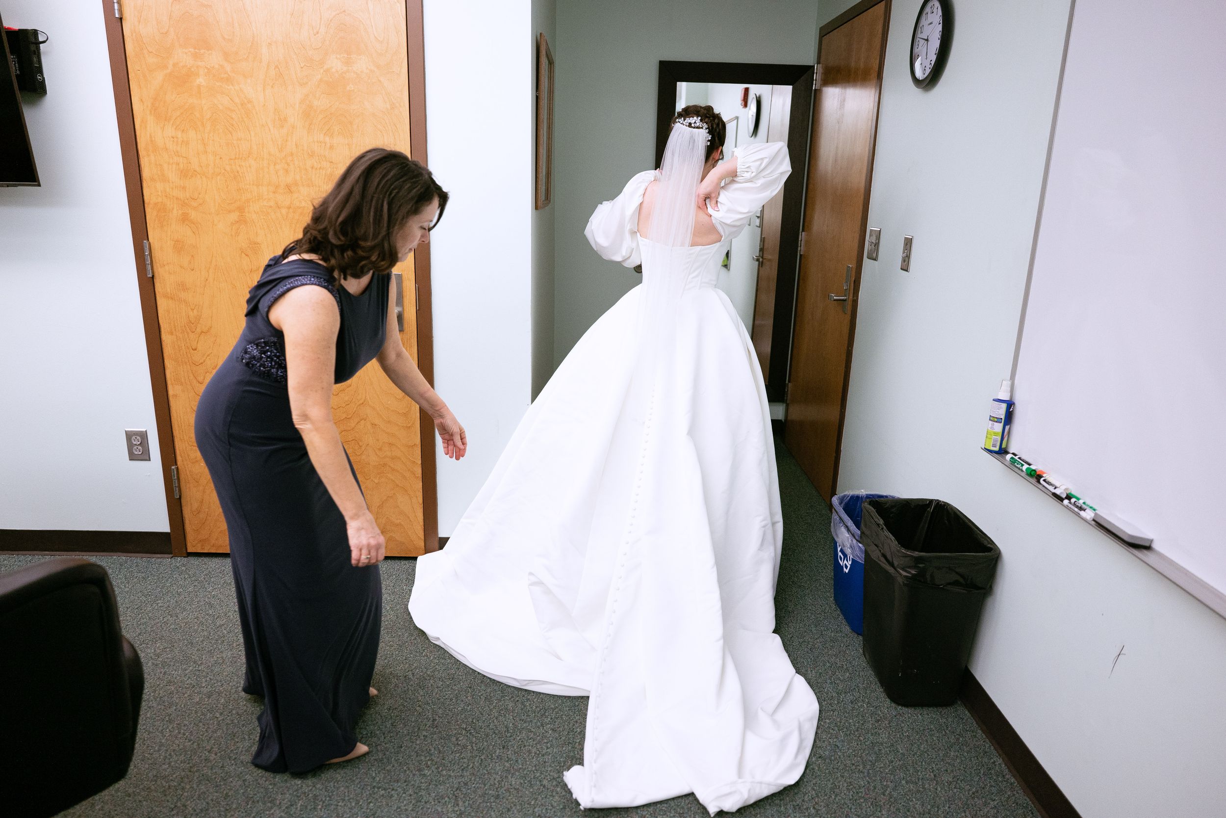 A person helps adjust a bride's white wedding dress while getting ready in a room.