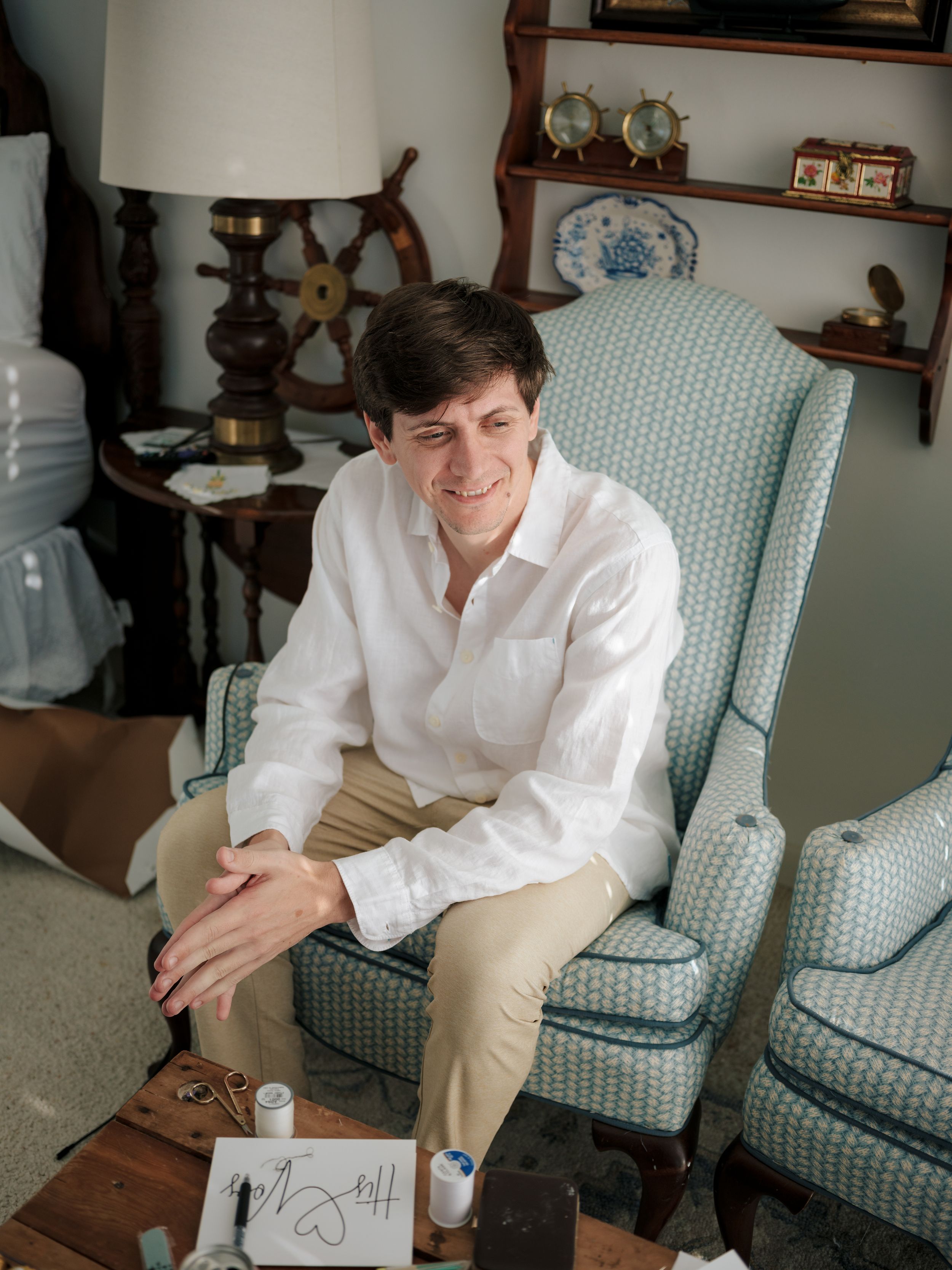 Person in white shirt and khaki pants sits in a blue patterned armchair writing something.