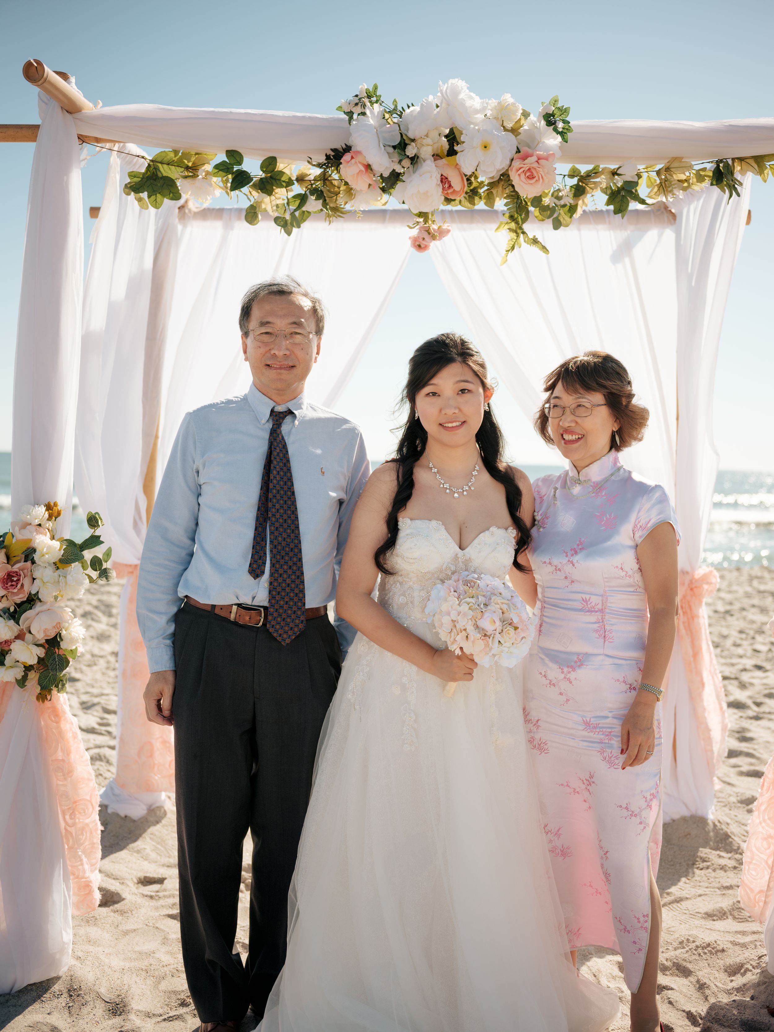 Wedding ceremony setup on beach with white draped arch and floral decorations.