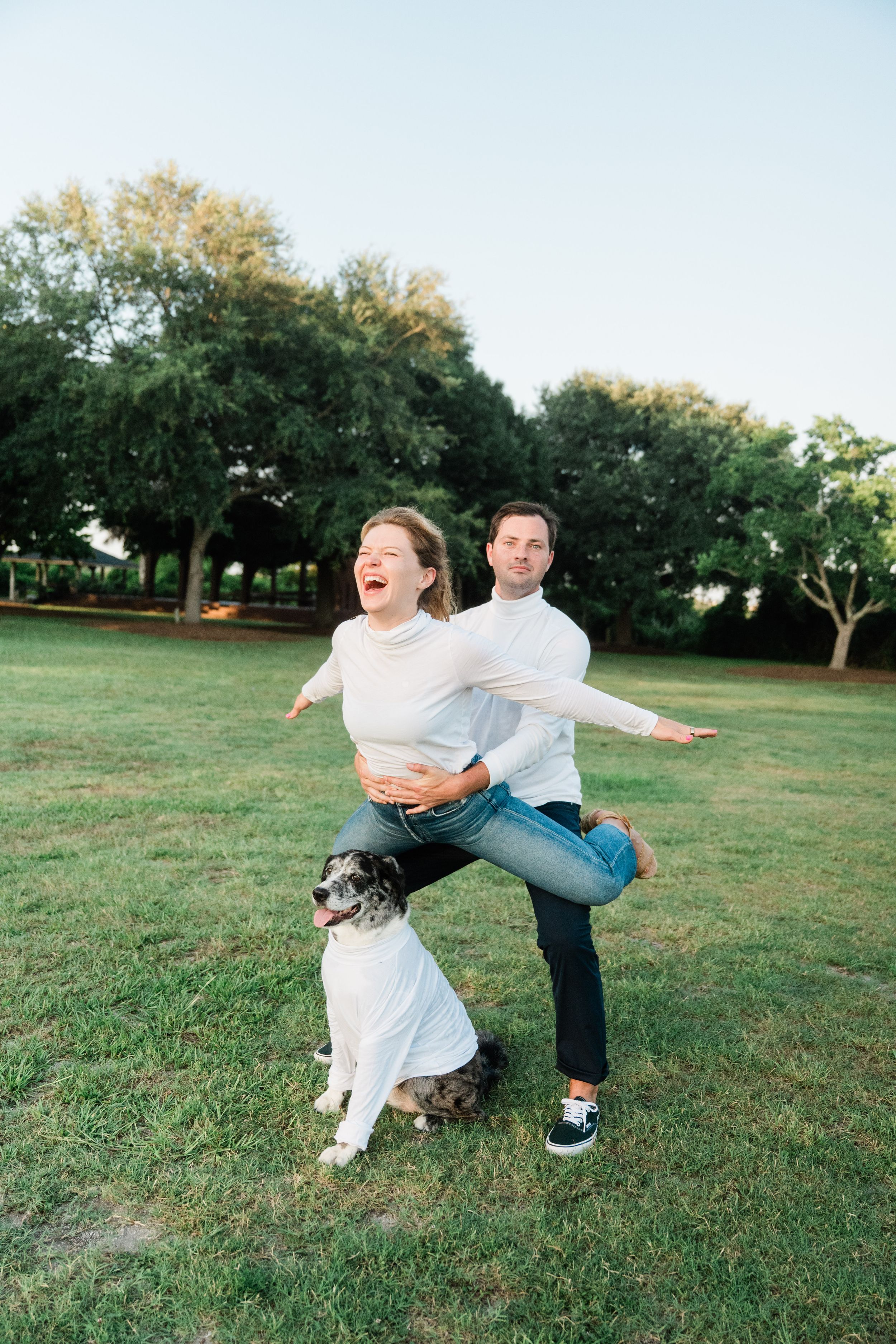 A playful couple dances together on a grassy lawn with trees in the background during golden hour.