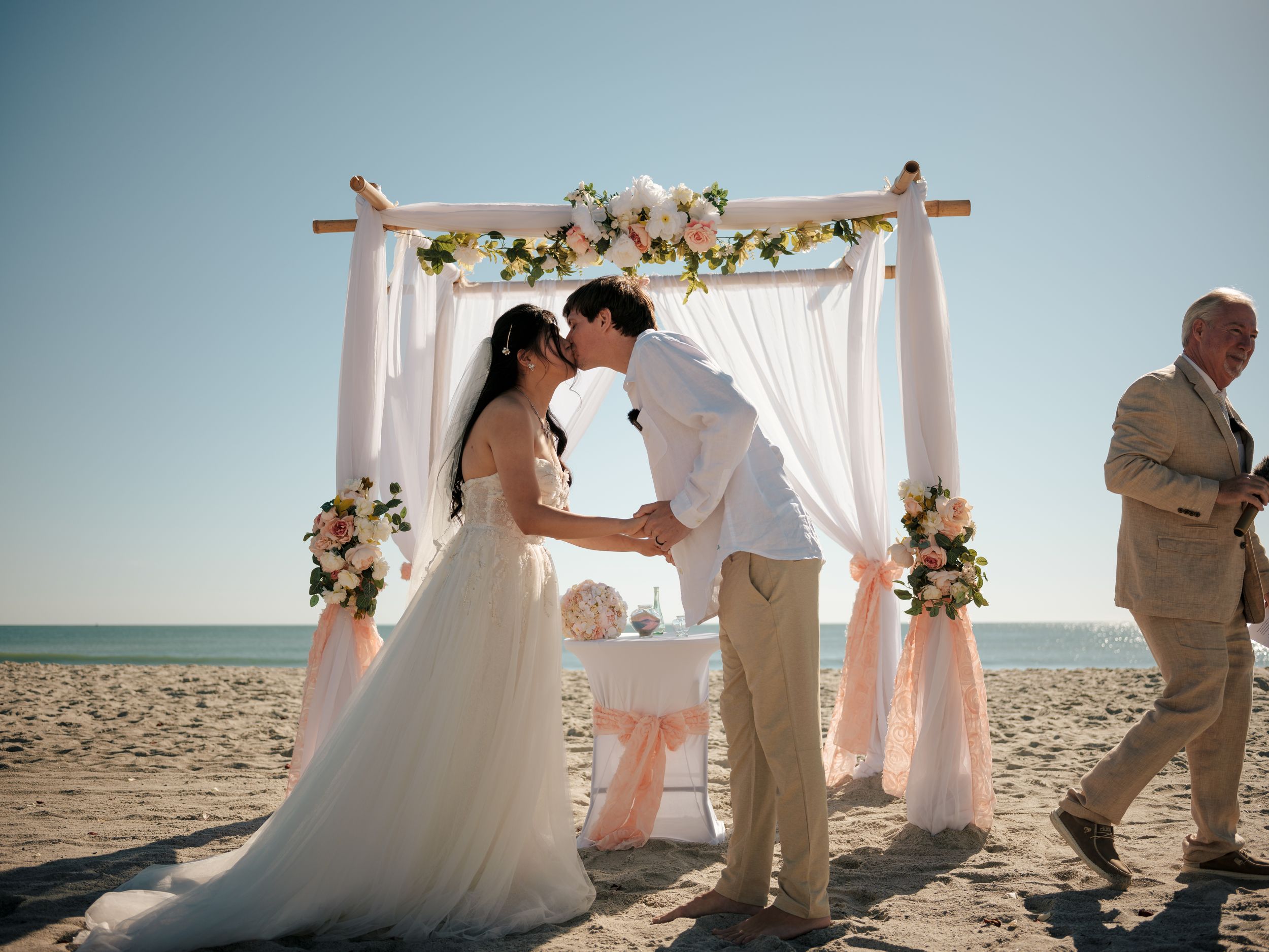Beautiful beach wedding ceremony under white arch decorated with flowers.