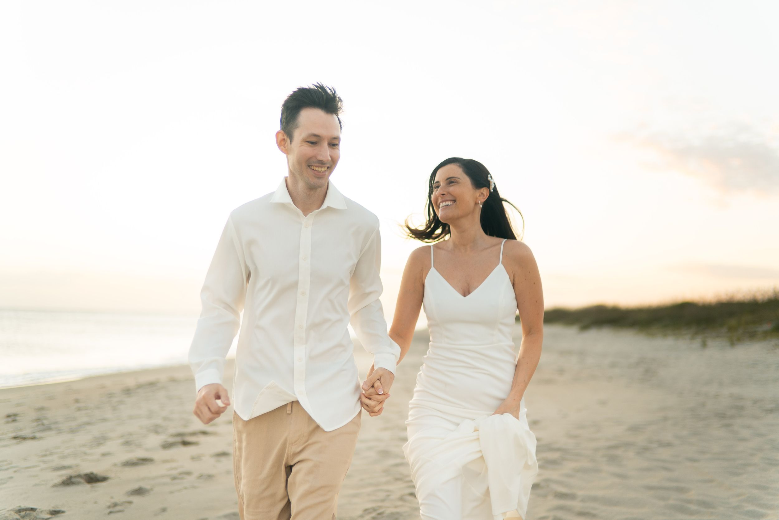 A couple in white attire walks hand in hand along a sandy beach at sunset, creating a romantic silhouette.