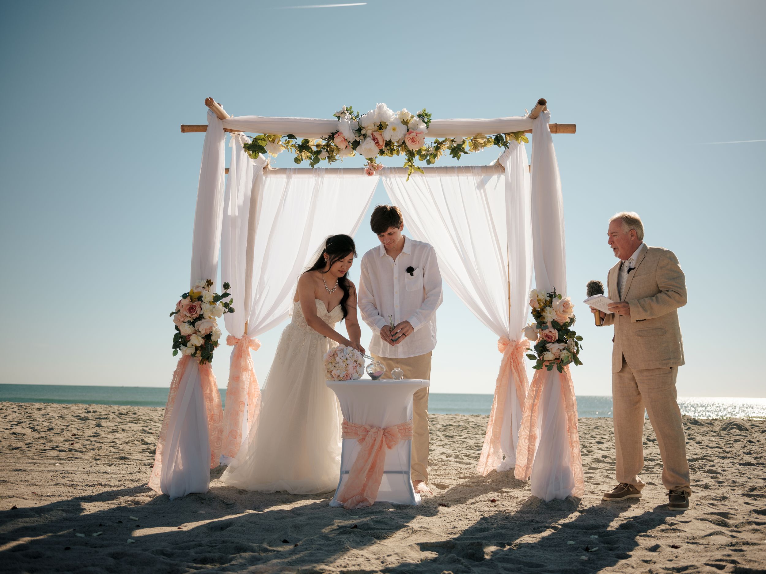 White wedding arch decorated with flowers stands on sunny beach during seaside ceremony.