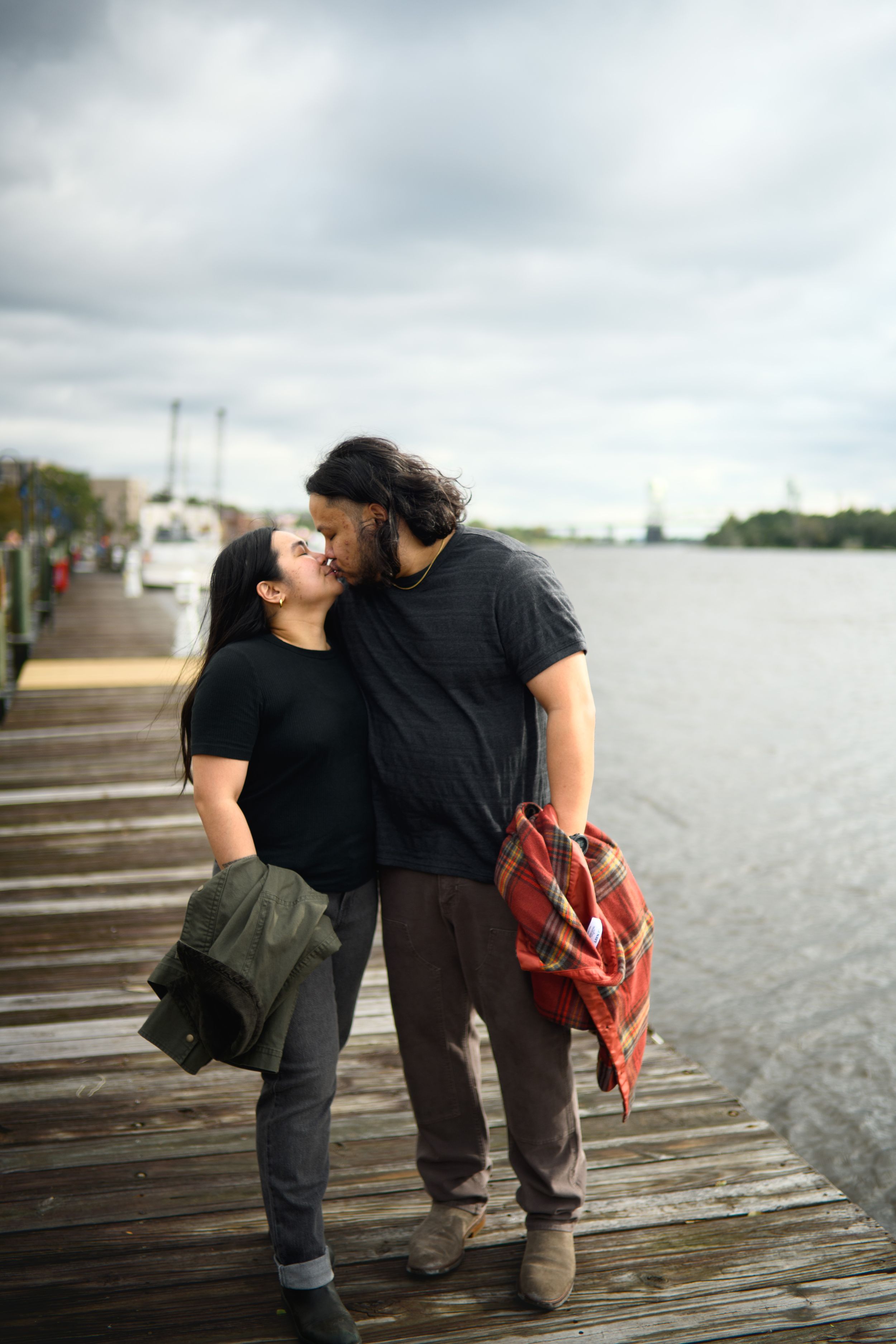 A couple in matching black shirts share an intimate moment on a wooden dock by the water.