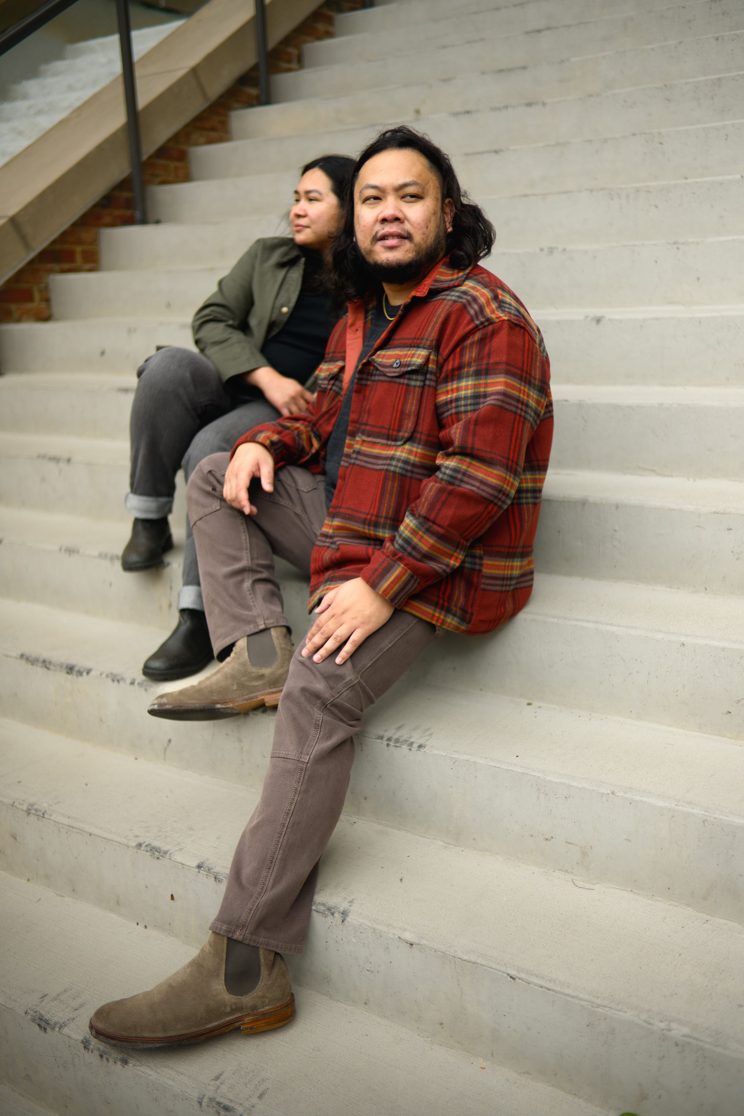 Two people sit casually on concrete steps wearing casual fall attire including plaid flannel and boots.