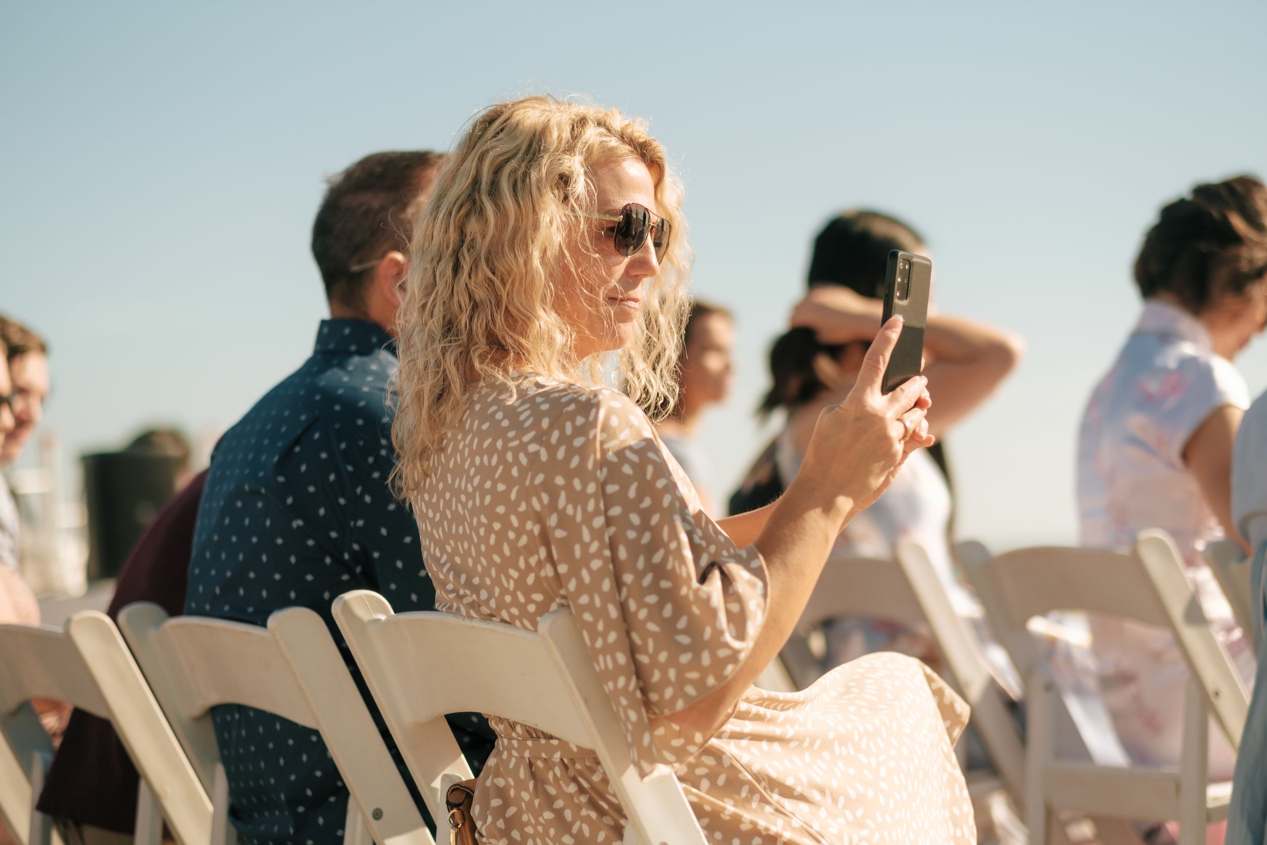 People seated in rows at an outdoor event with blue sky in background.