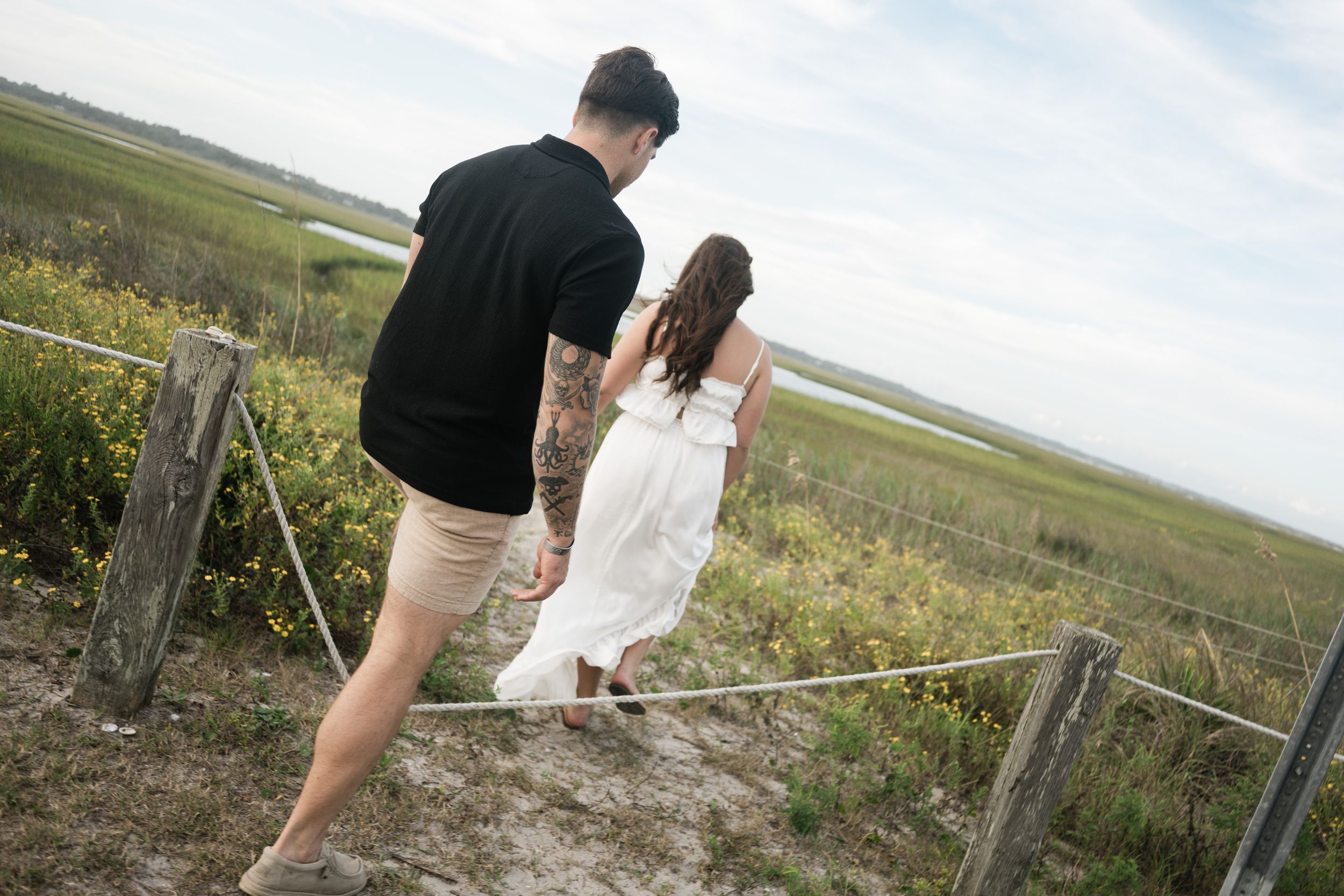 Couple walking along a scenic coastal boardwalk path surrounded by marsh grass and water views.