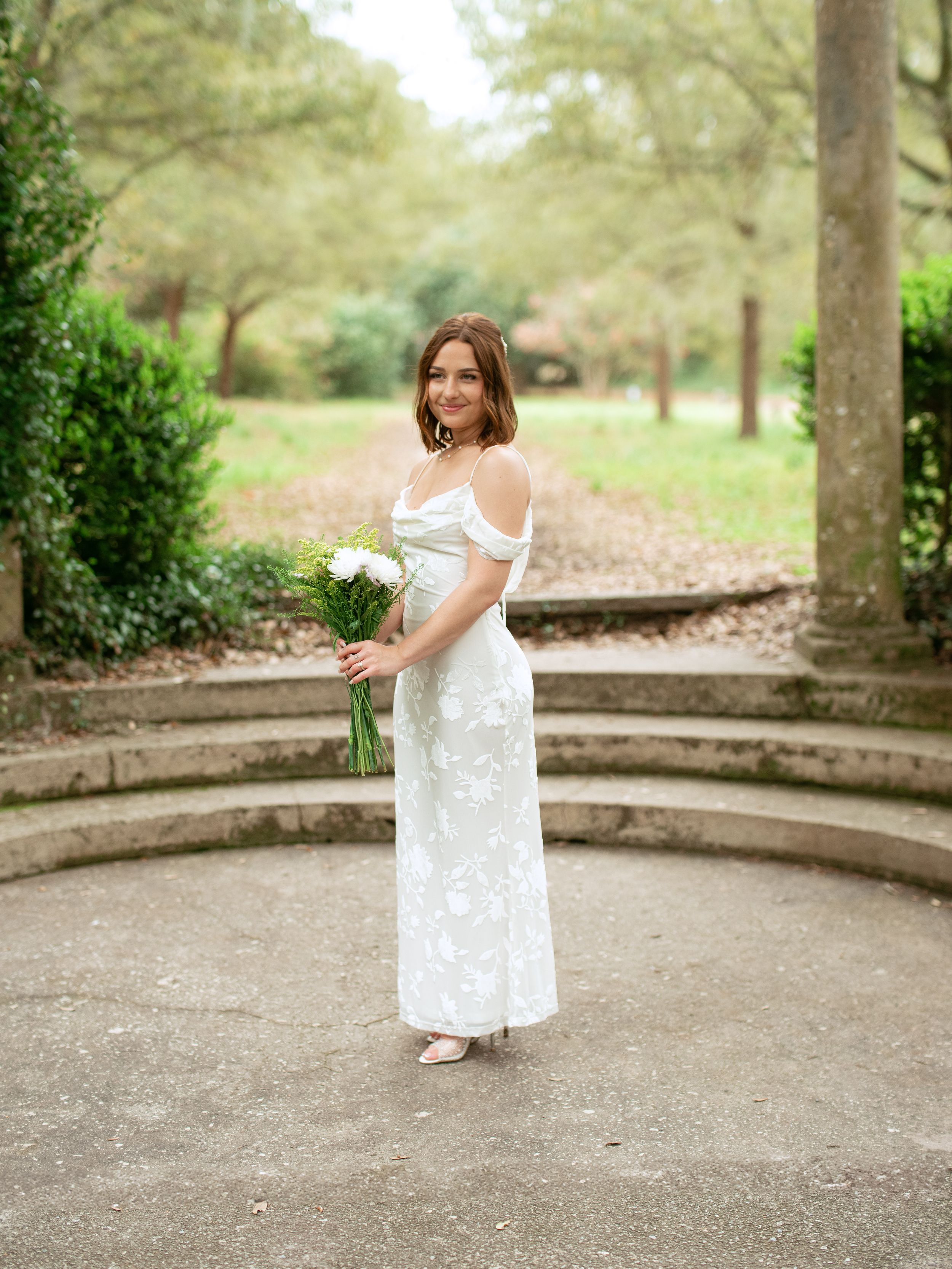 A bride in an elegant white off-shoulder dress poses with a bouquet on circular stone steps in a garden setting.