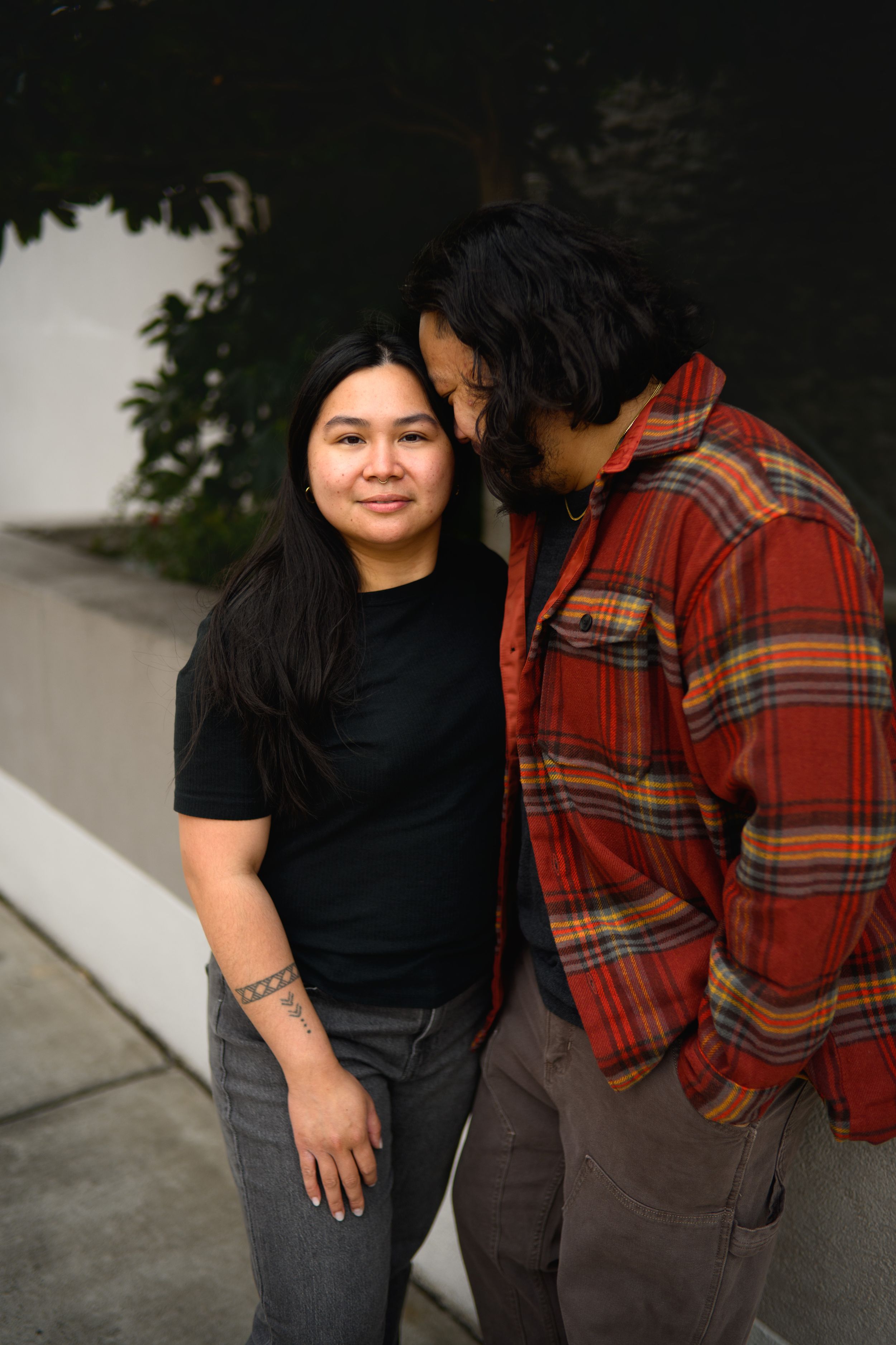 Two people in casual clothing, one wearing a black shirt and one in a plaid flannel, pose together outdoors.