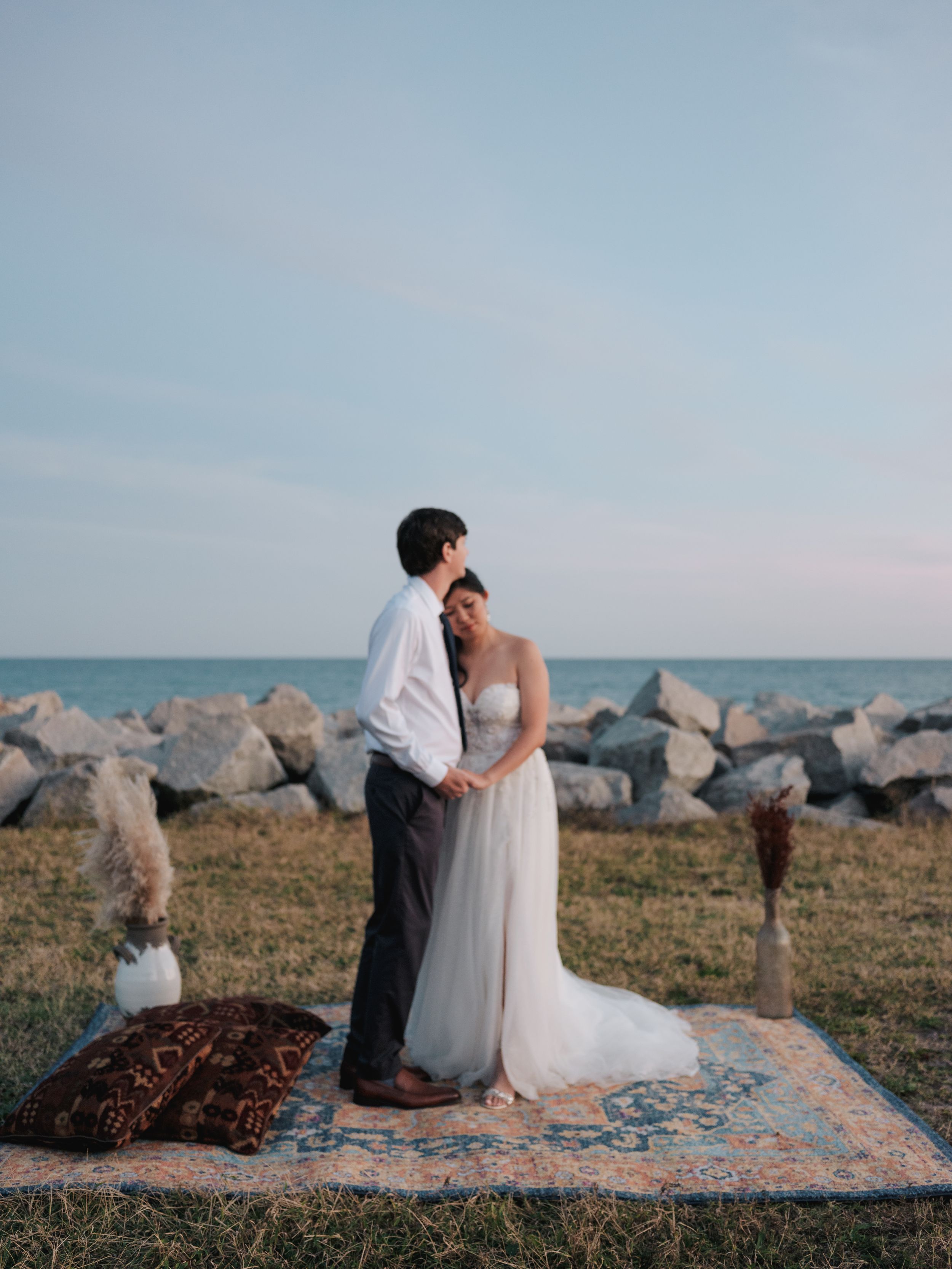 A couple in wedding attire sits beside the ocean on rocky shores during sunset.
