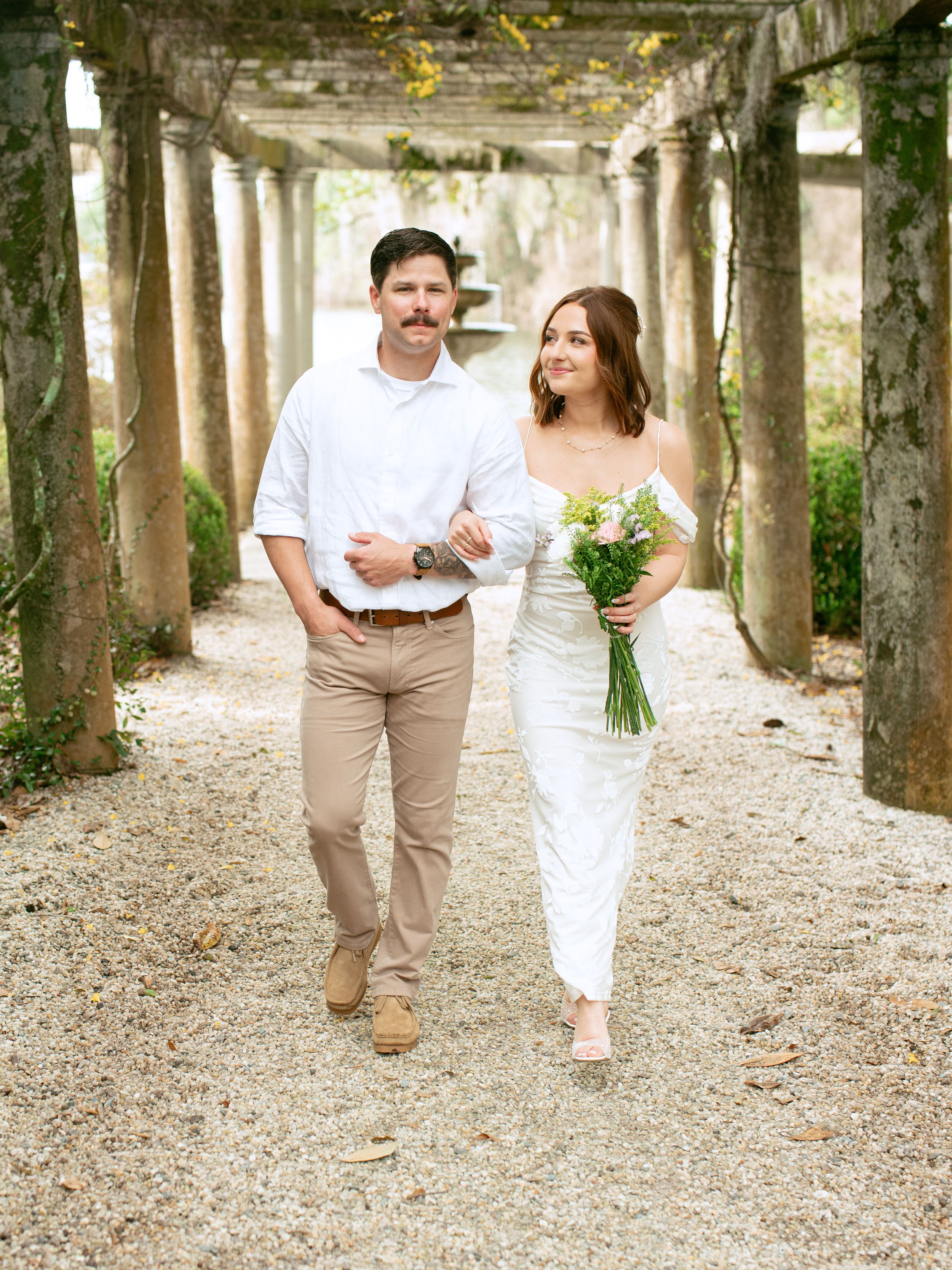 A couple walks together under a covered garden path pergola, with the bride holding a bouquet in a simple white dress.