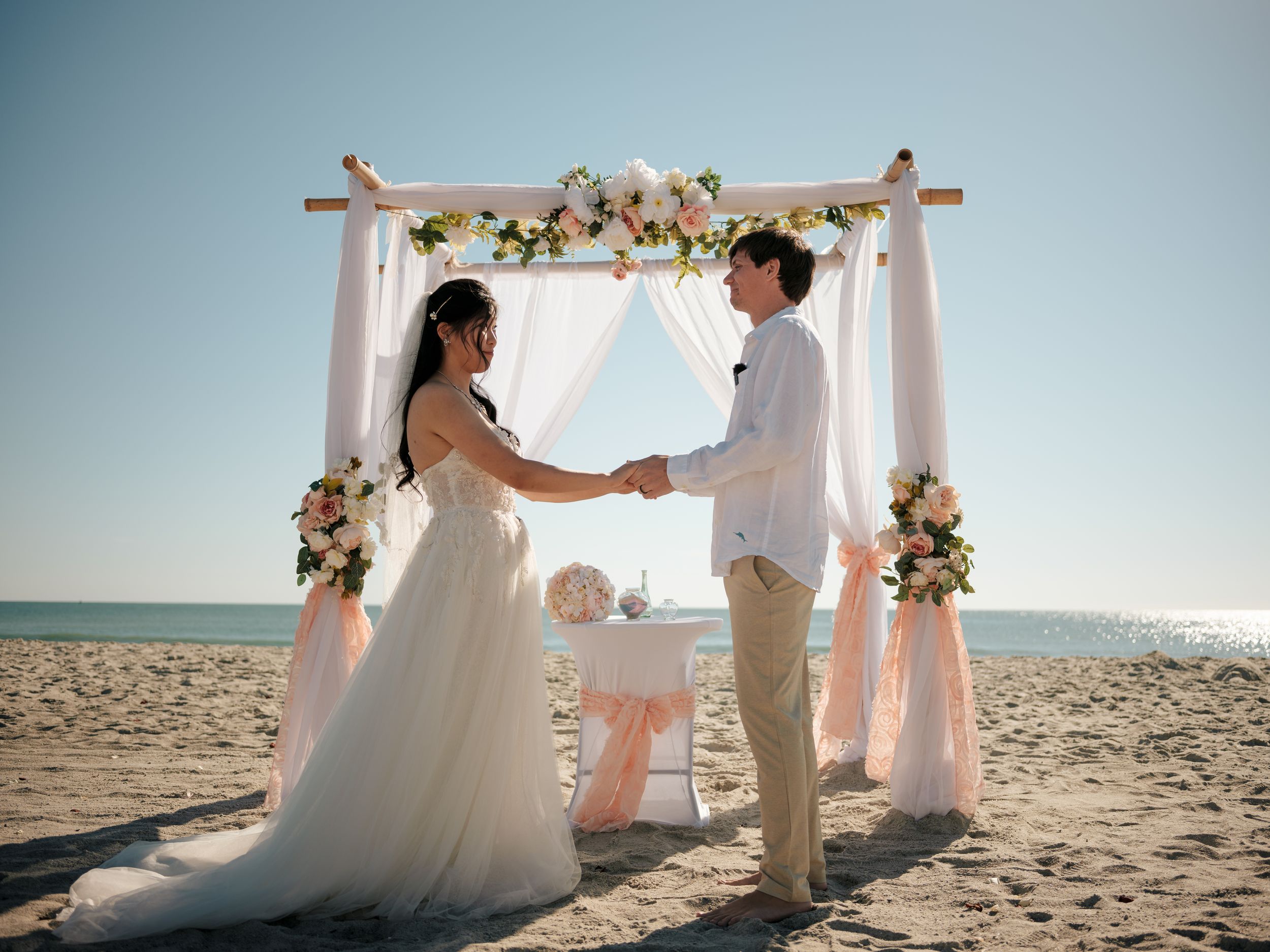 Couple exchanges vows under floral arch during beachside wedding ceremony.