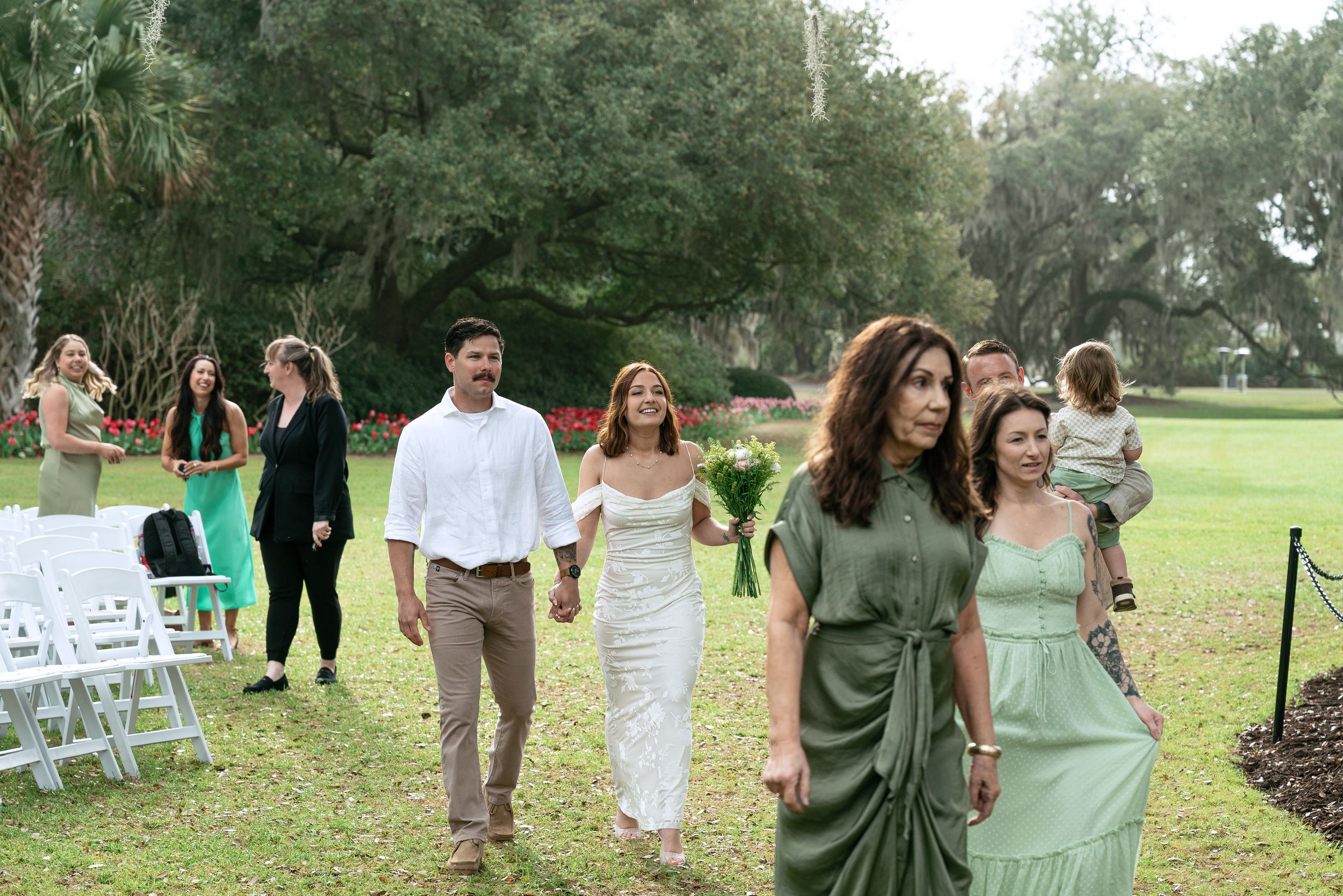 Wedding guests in formal attire walking through a lush garden setting with white chairs arranged for an outdoor ceremony.