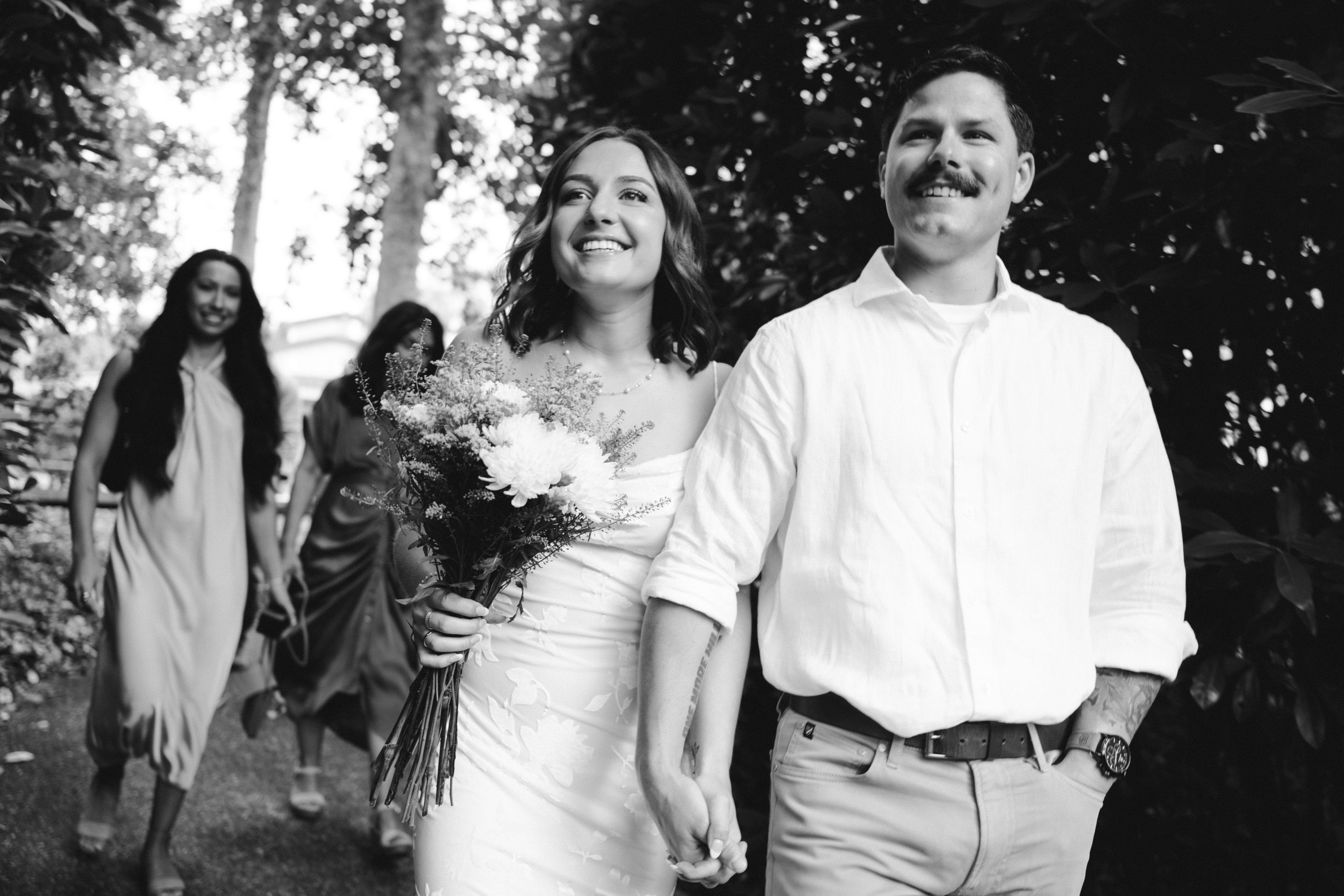 A black and white wedding portrait showing a couple walking together with a bouquet while guests follow in the background.