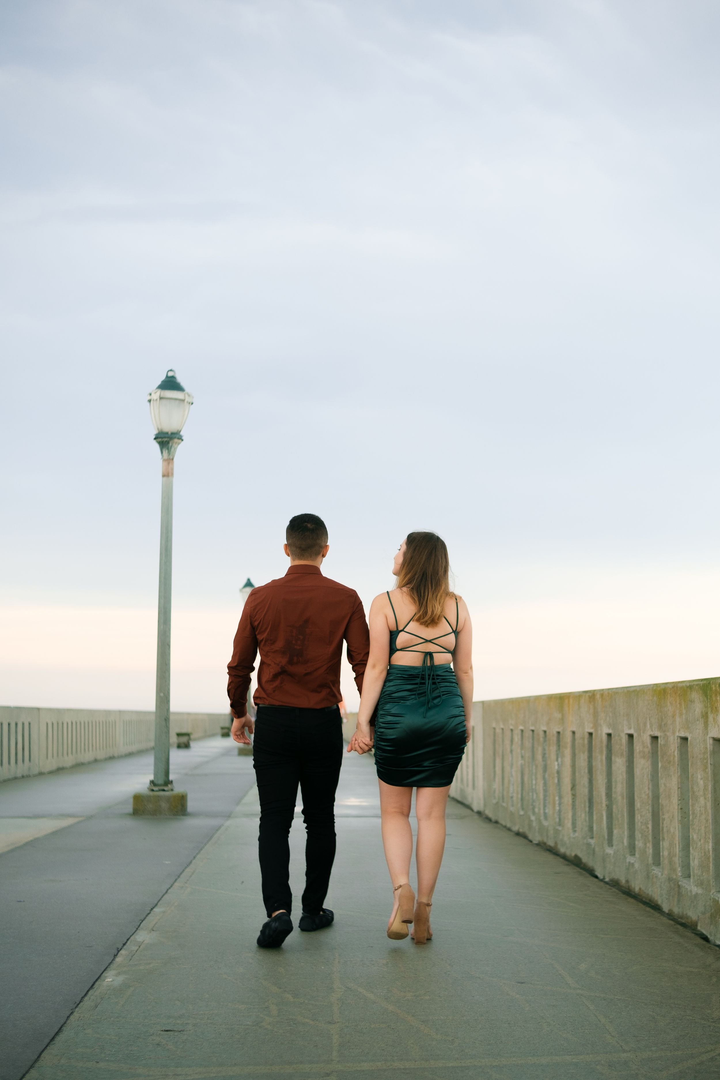Two people walk hand in hand along a concrete pier with lampposts and ocean views.