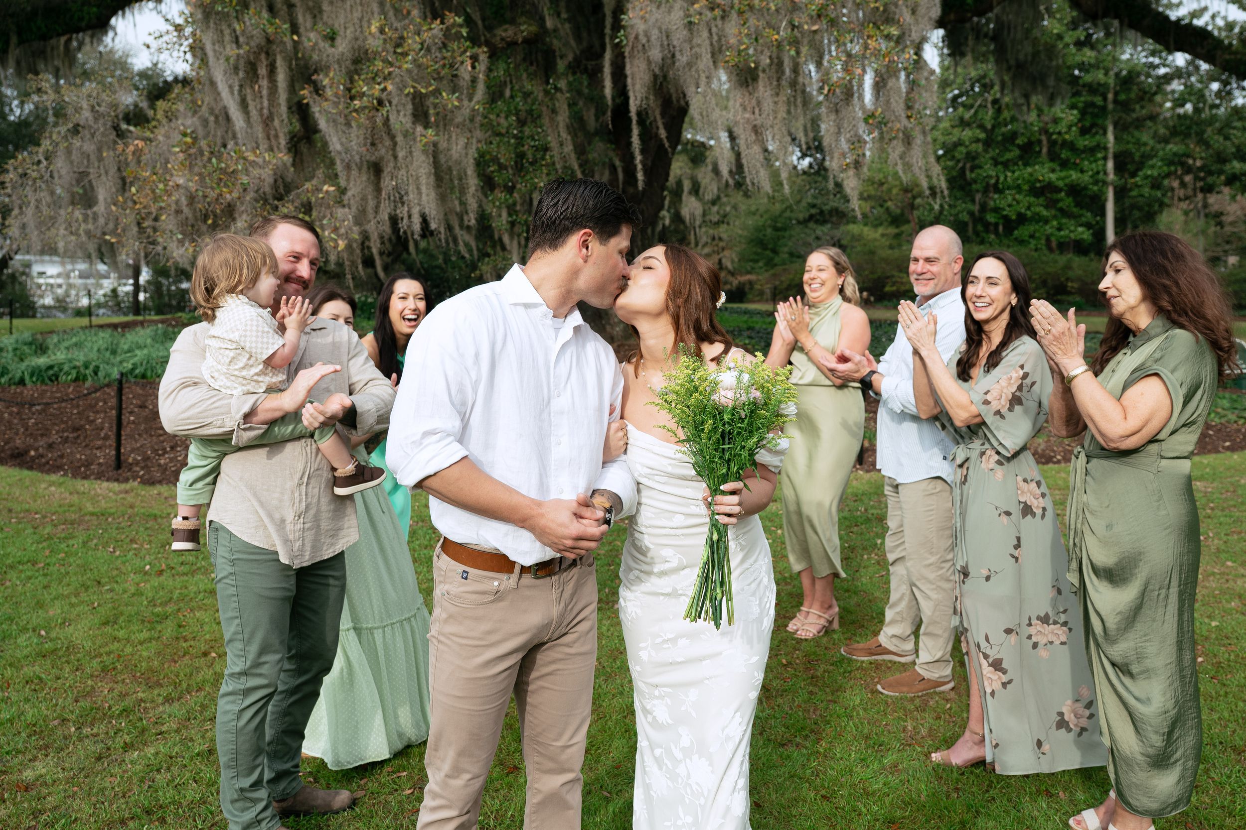 Wedding party wearing khaki and sage green attire celebrates together outdoors under moss-draped trees.
