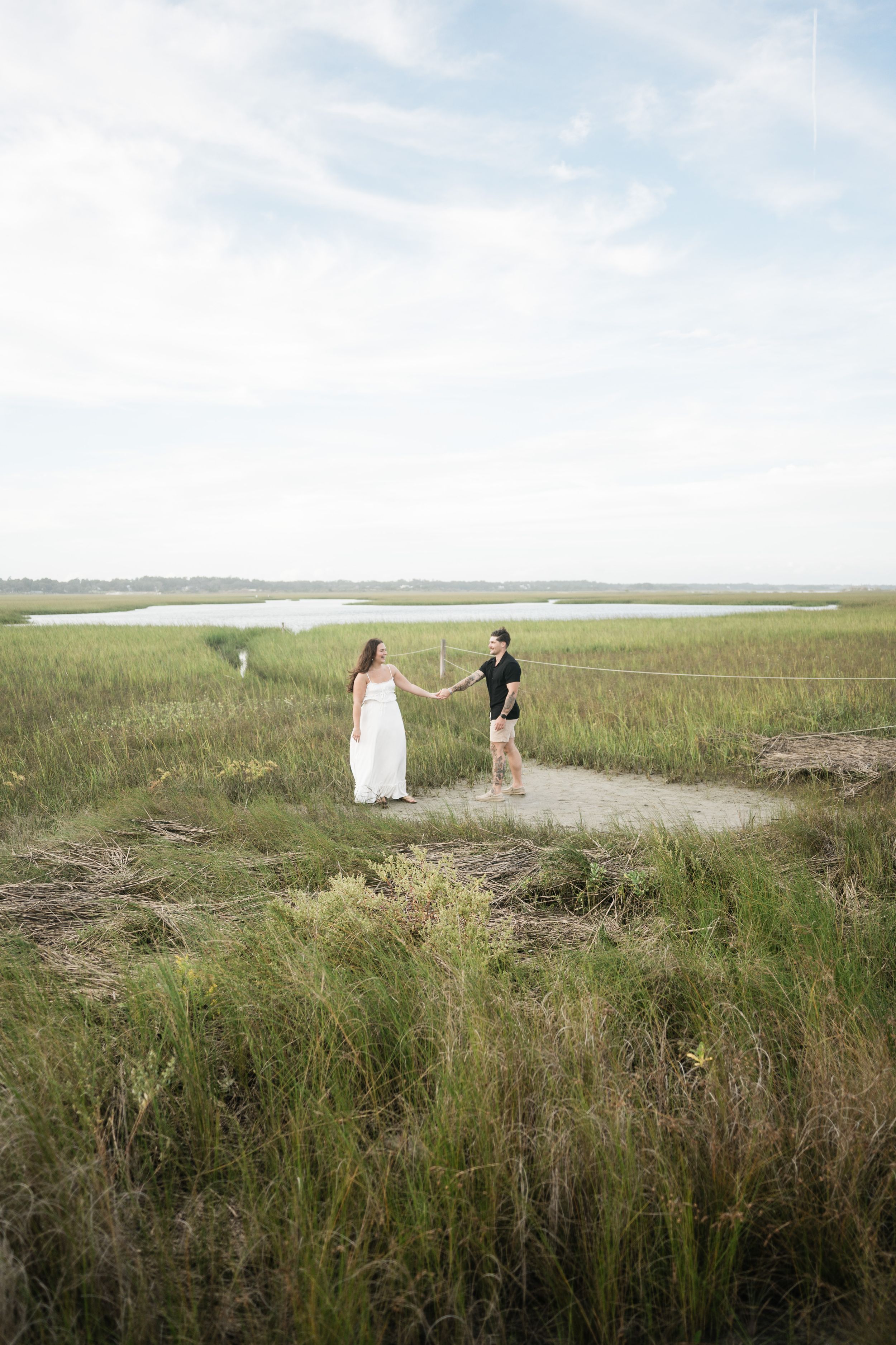 A couple walks along a scenic coastal path with tall grass and water views in the background.