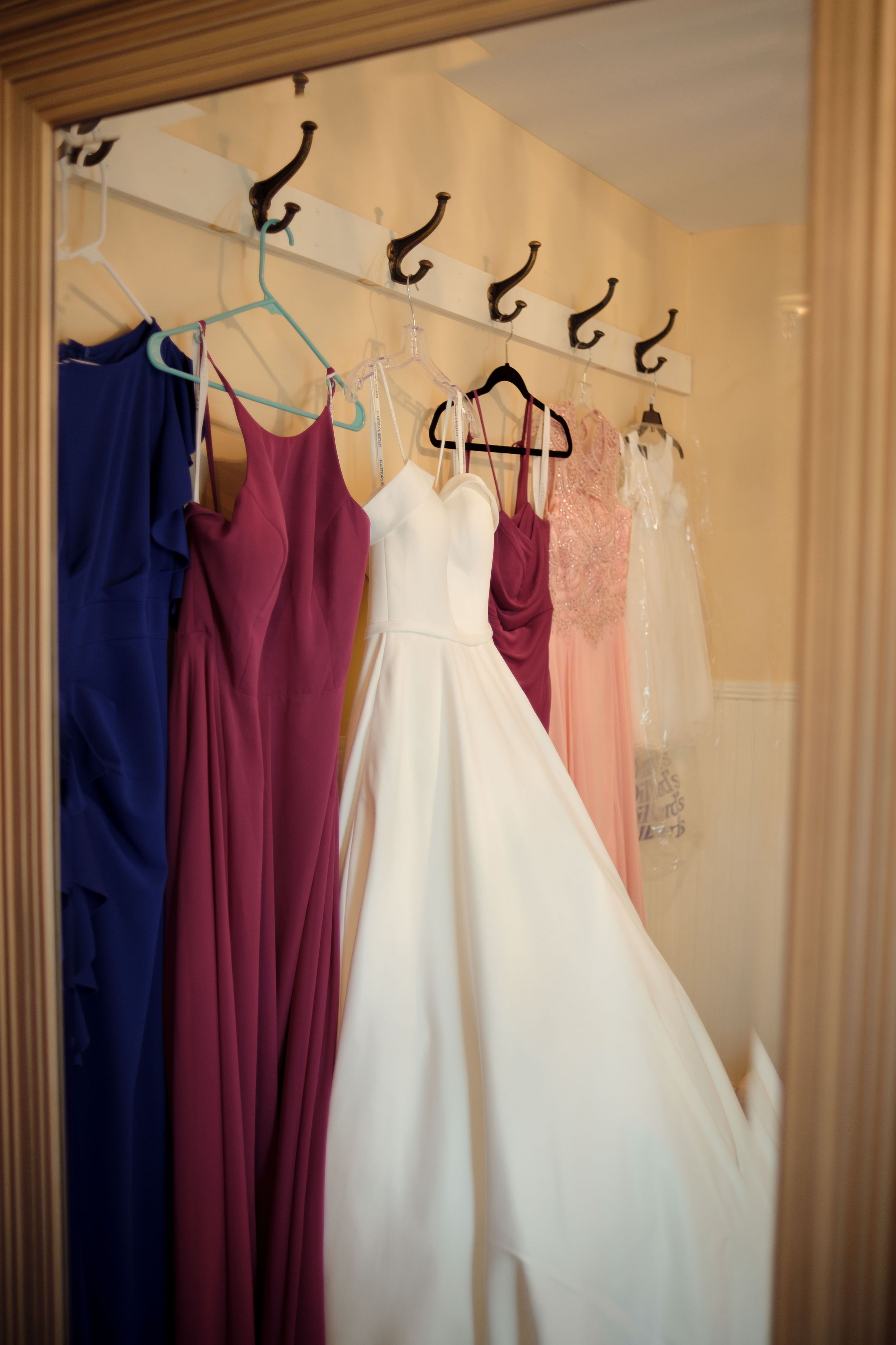 Wedding gowns and bridesmaid dresses in navy and burgundy hang neatly on wooden hangers against a cream-colored wall.
