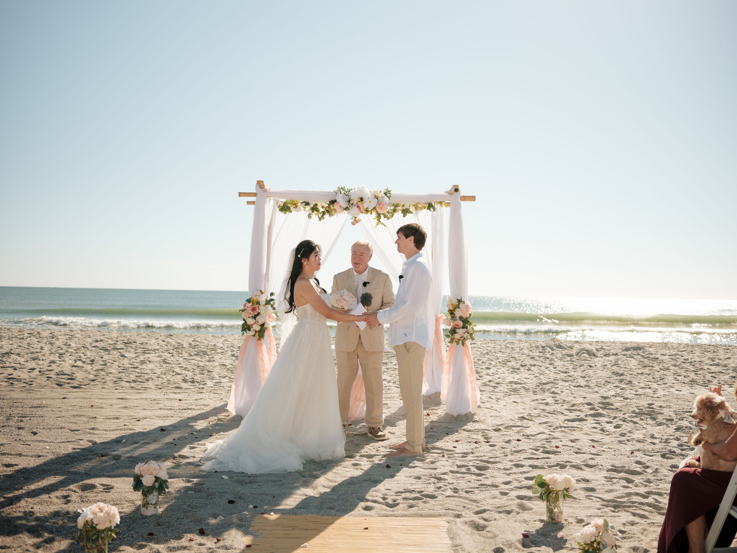 A beachside wedding ceremony setup with white floral arch and chairs on the sand.