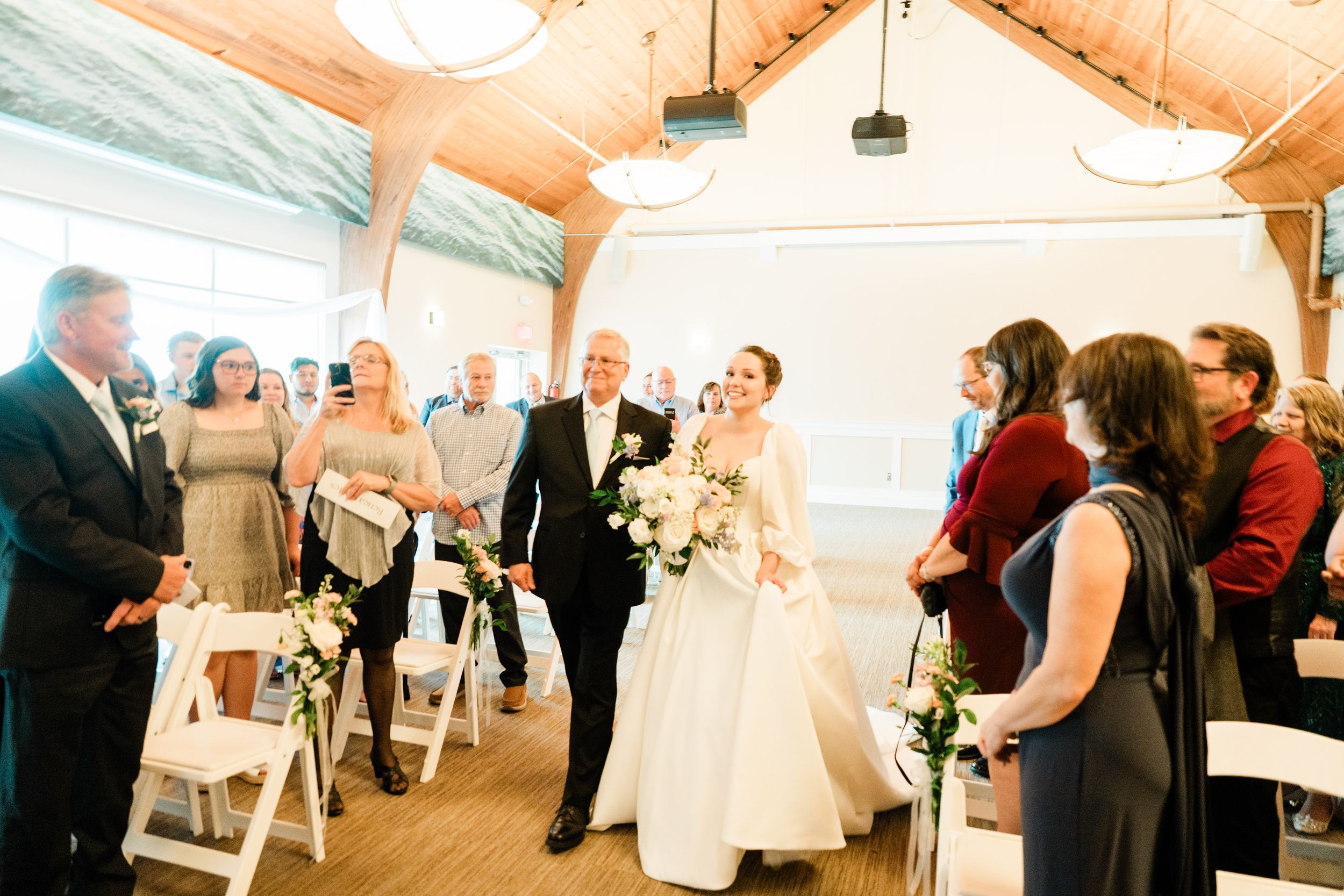 Wedding ceremony guests gather in a bright indoor venue with wooden beams and white chairs.