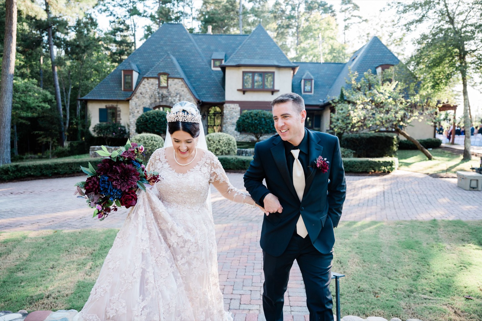 A couple in wedding attire walks along a brick pathway in front of a charming blue-roofed house.