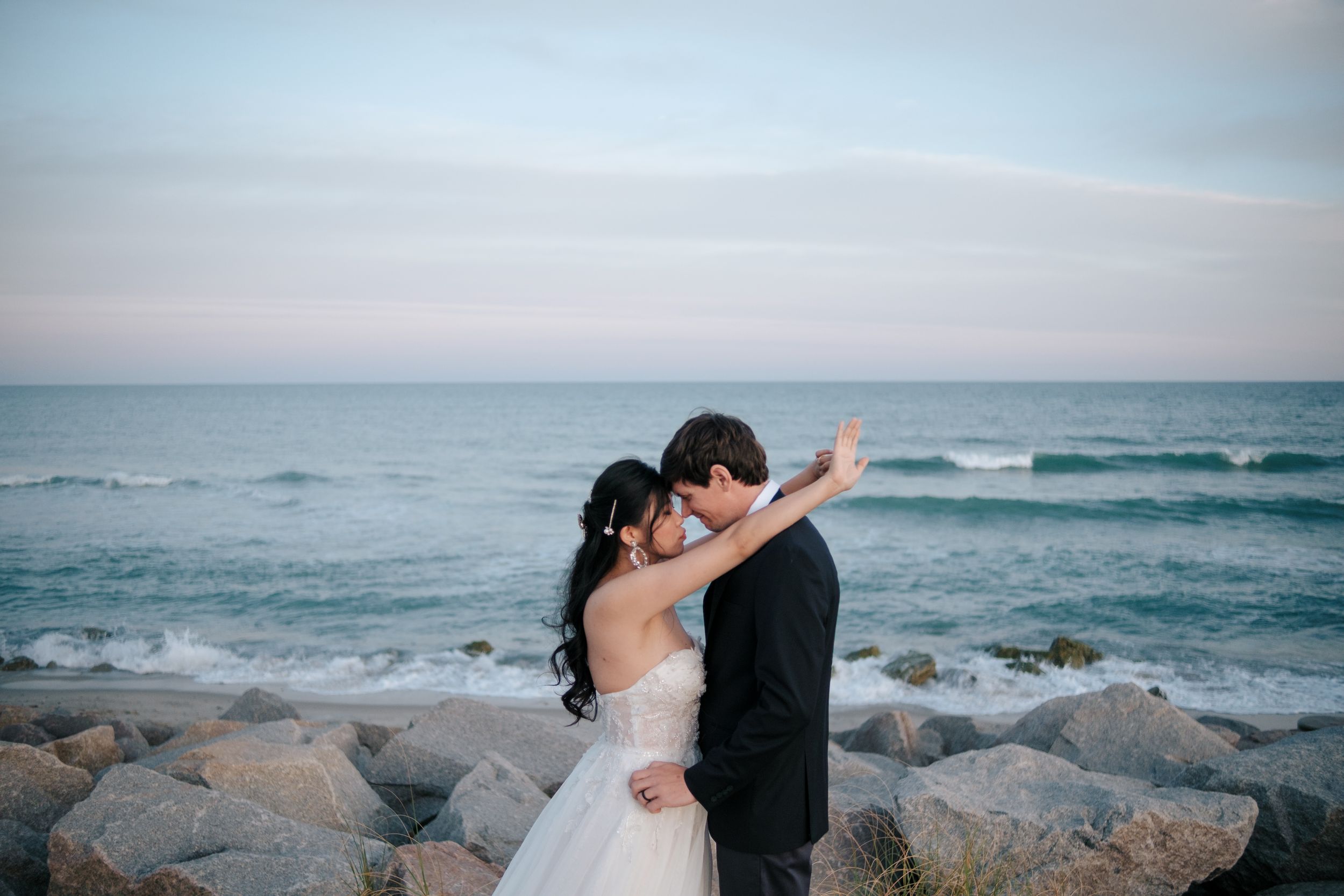A couple shares an intimate moment against a rocky beach background as waves crash on the shore at sunset.