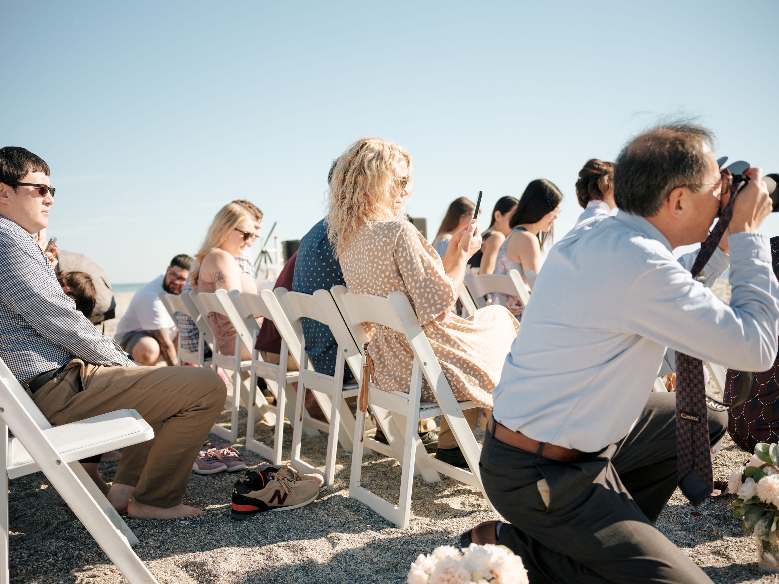 Wedding guests seated in white chairs on the beach during an outdoor ceremony.
