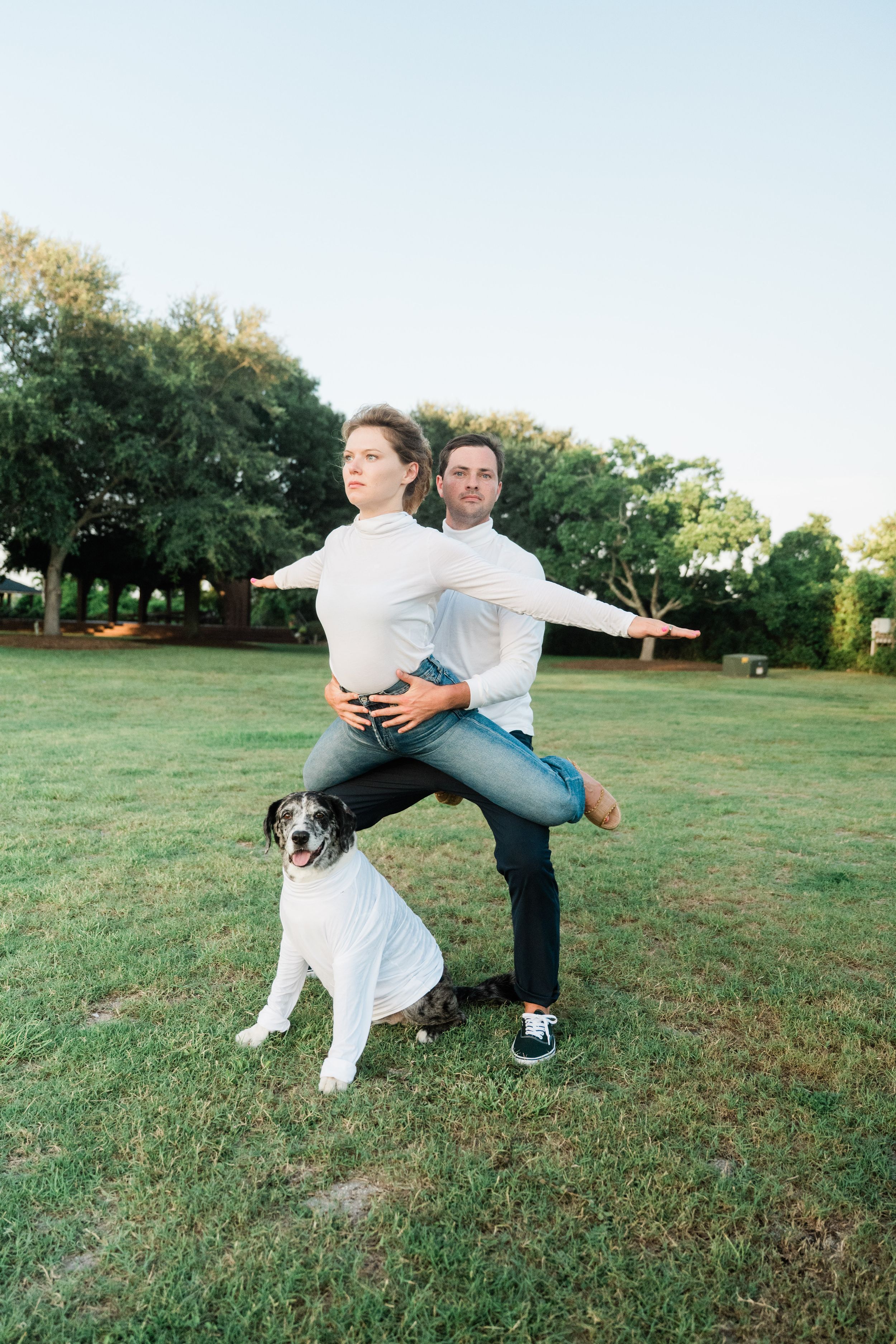 A playful couple poses with their white dog in a grassy park setting during a casual outdoor photoshoot.