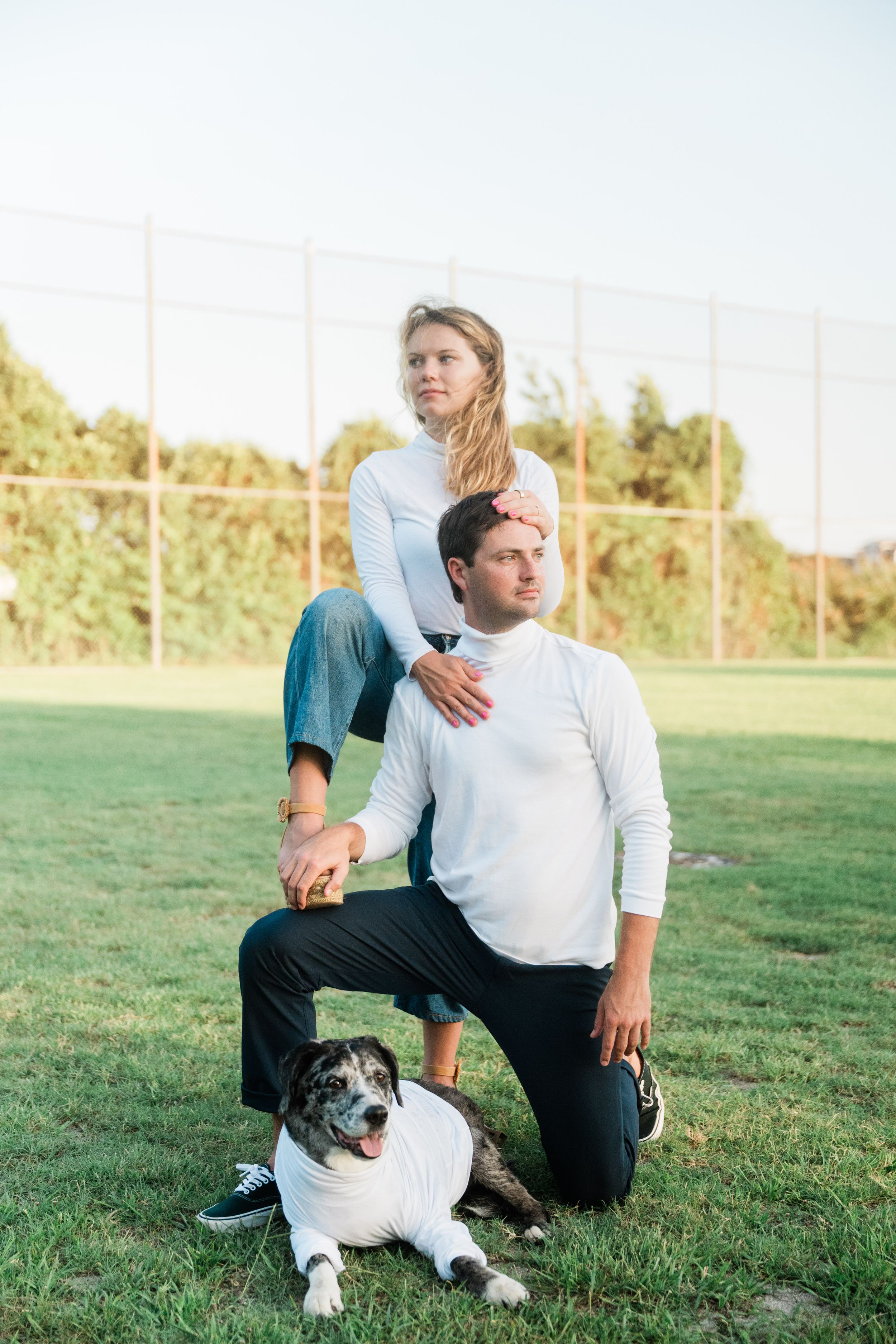 A couple poses outdoors on a grassy field with their black and white dog on a sunny day.