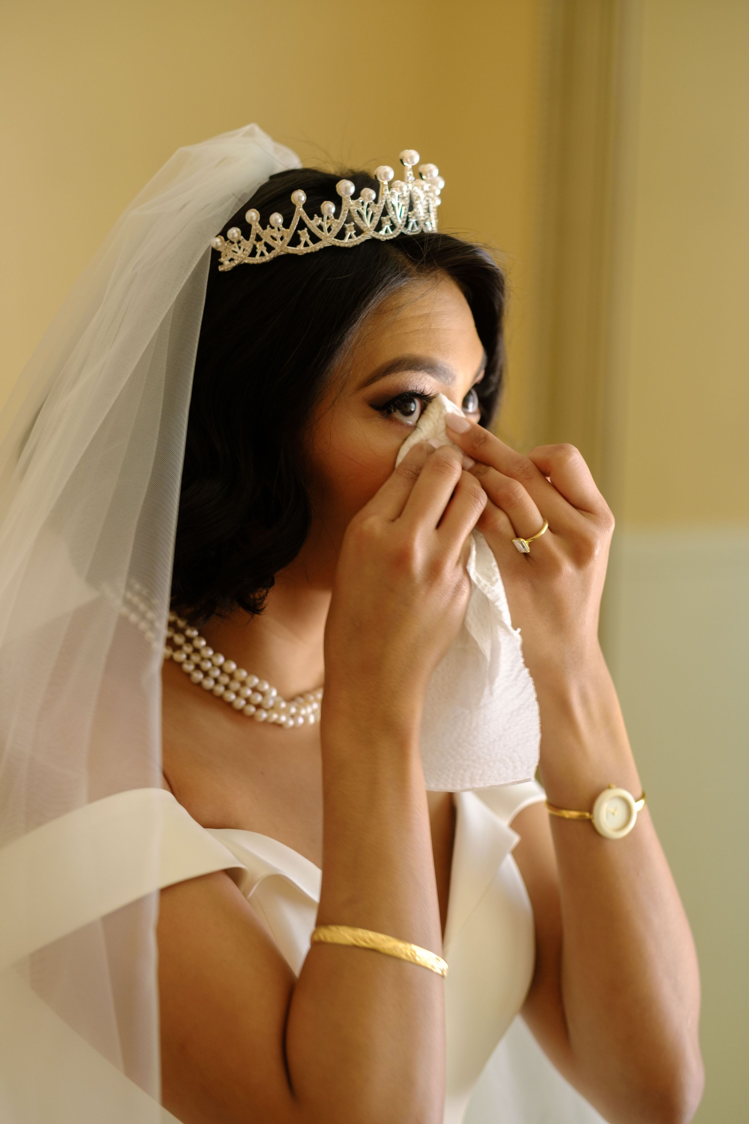 Wedding preparation scene showing someone dabbing tears with a tissue while wearing a delicate veil and pearl necklace.