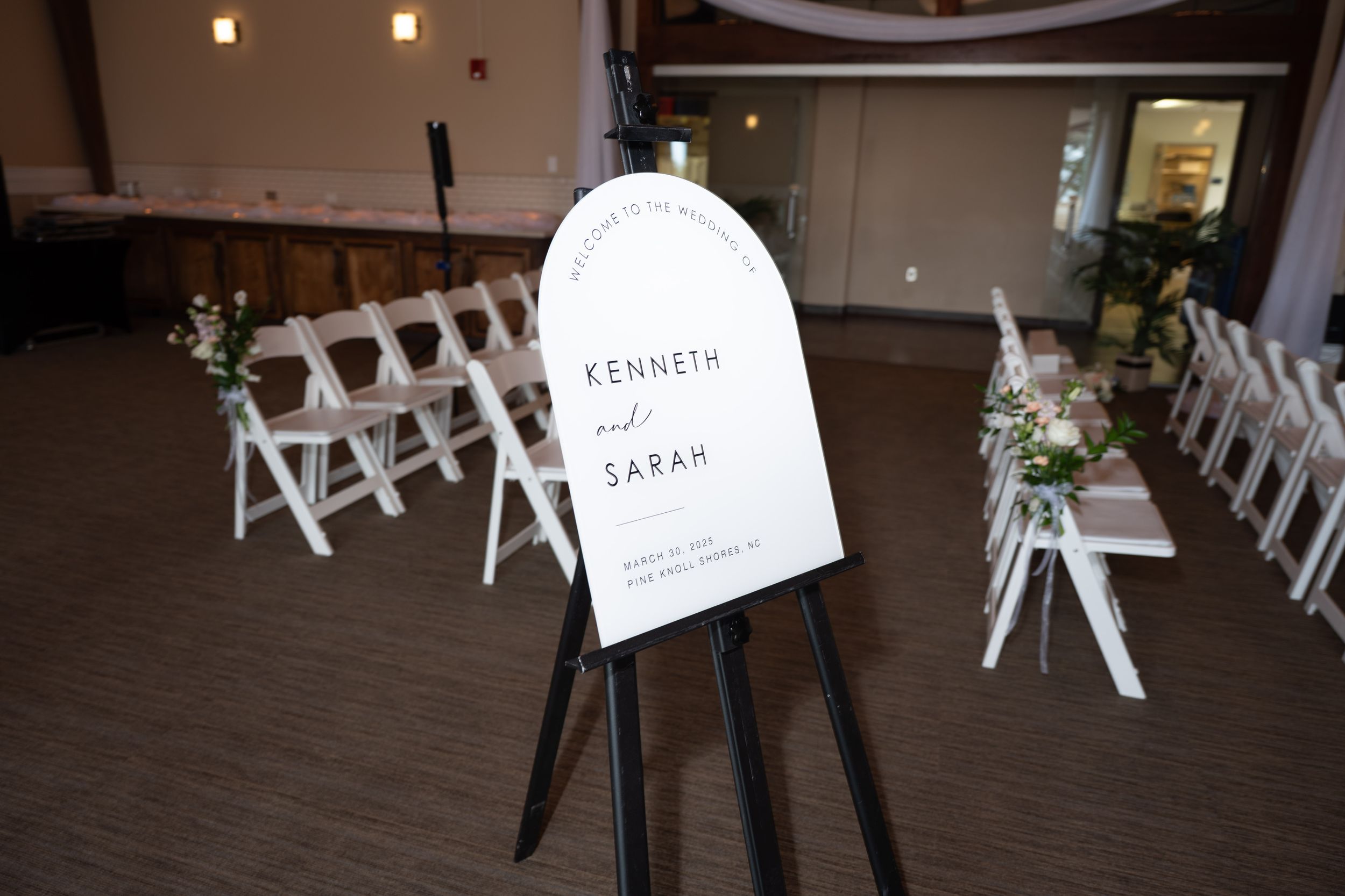 A wedding welcome sign on an easel stands in front of white folding chairs in an indoor venue.