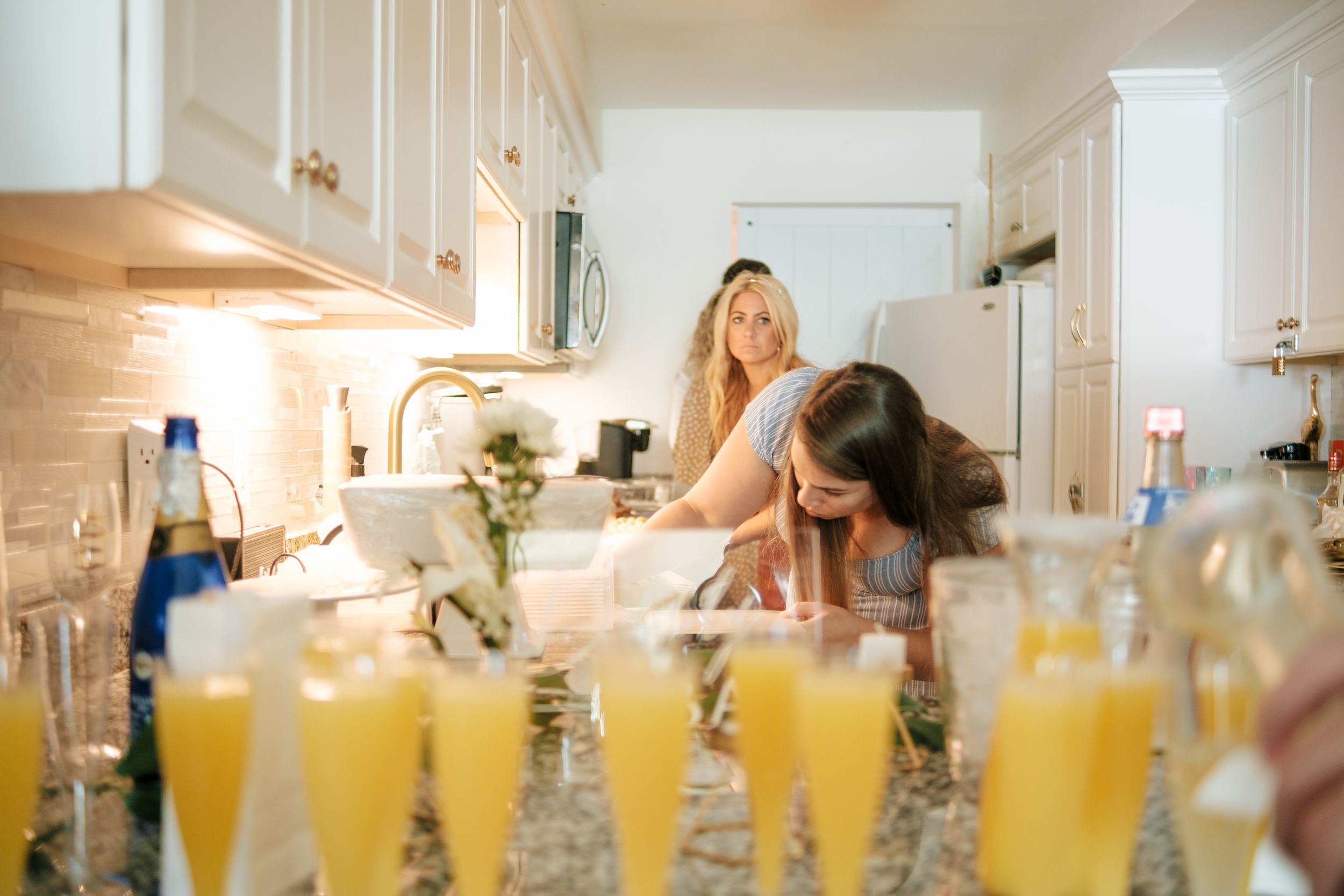 A row of mimosa cocktails in tall glasses lines a white kitchen counter during a gathering.